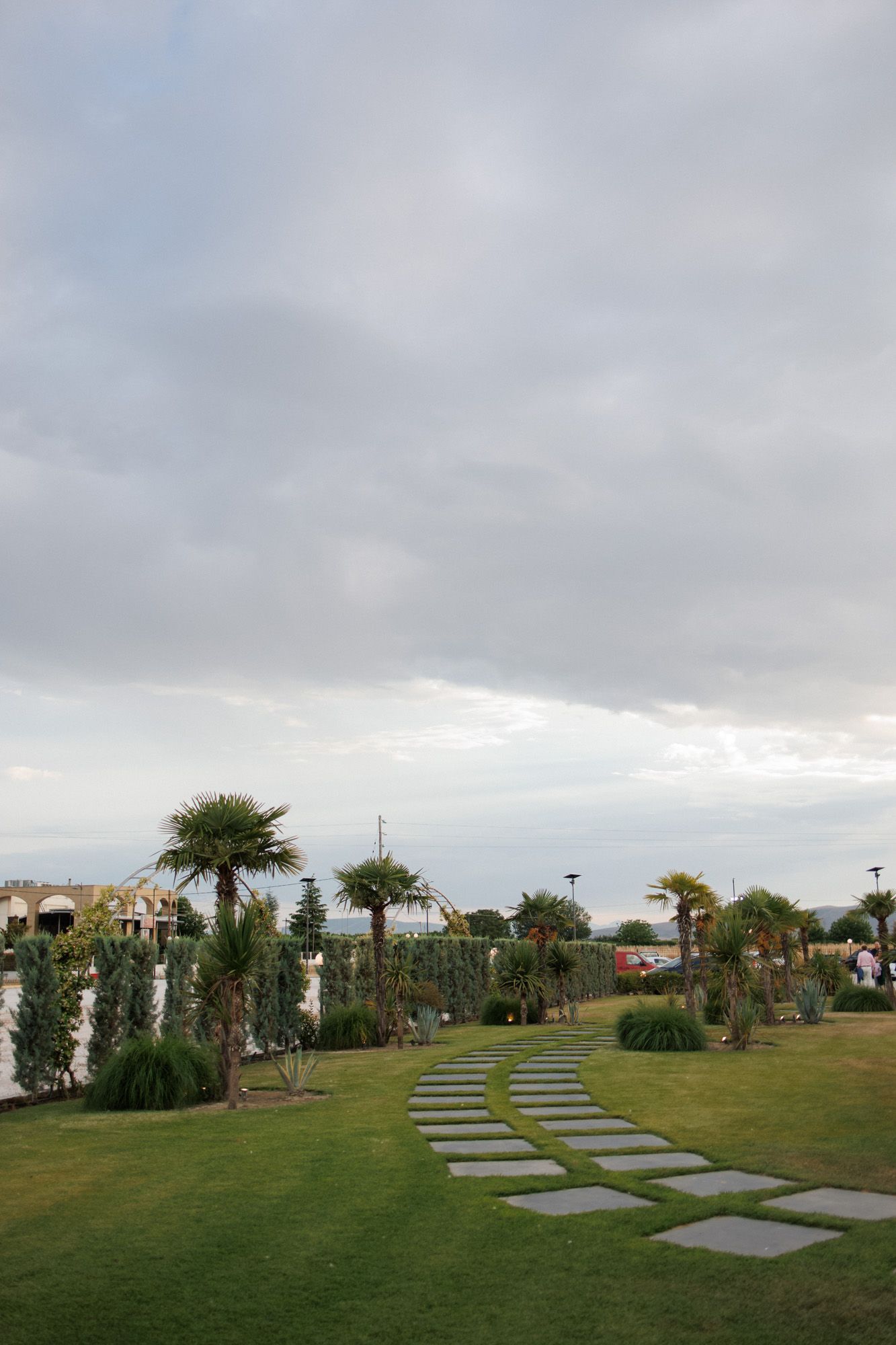 Lush garden path with stepping stones, palm trees, and manicured lawn under a cloudy sky.