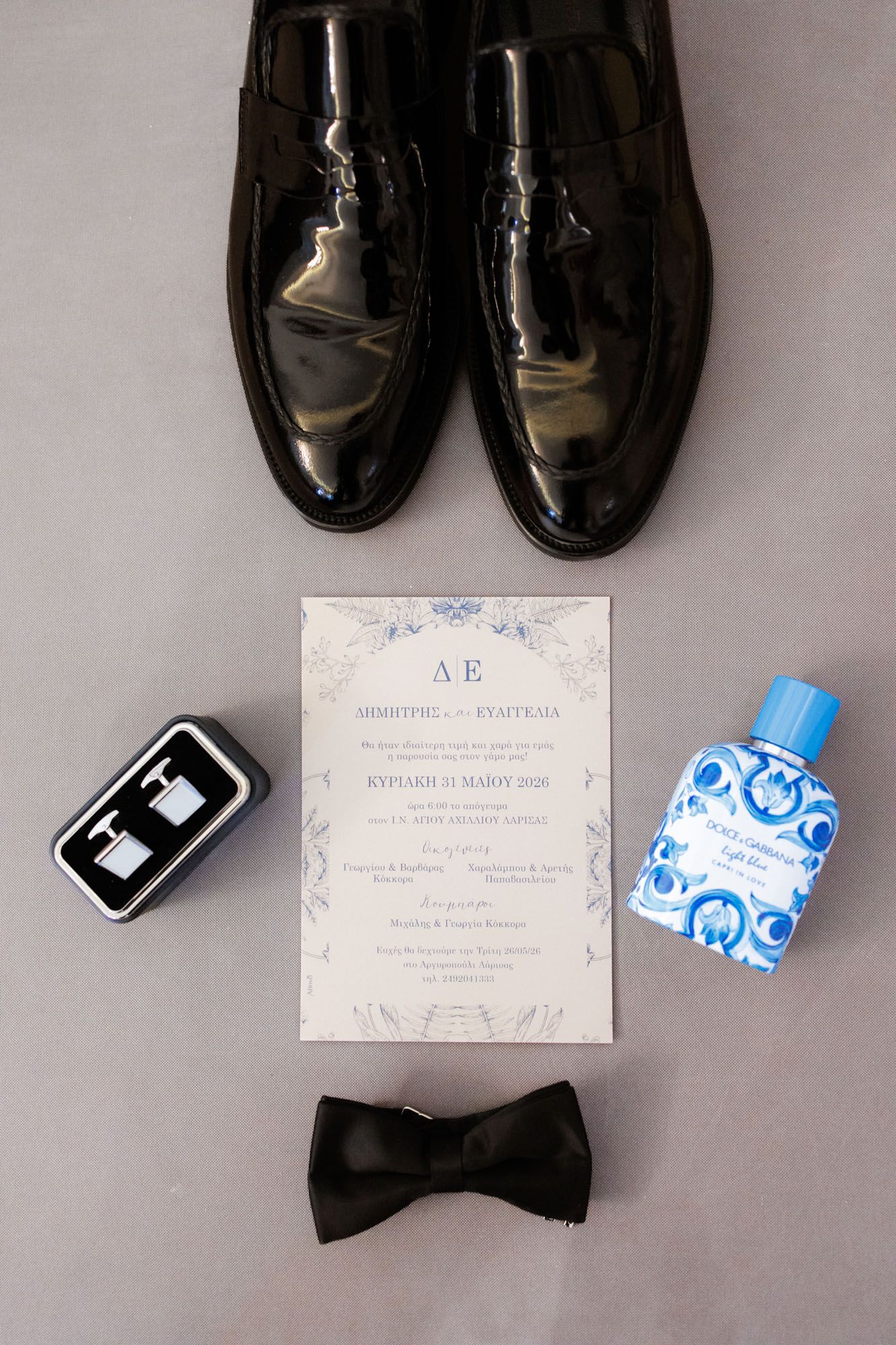 Groom wedding accessories flat lay with black patent leather shoes, bow tie, cufflinks, cologne, and elegant invitation card.