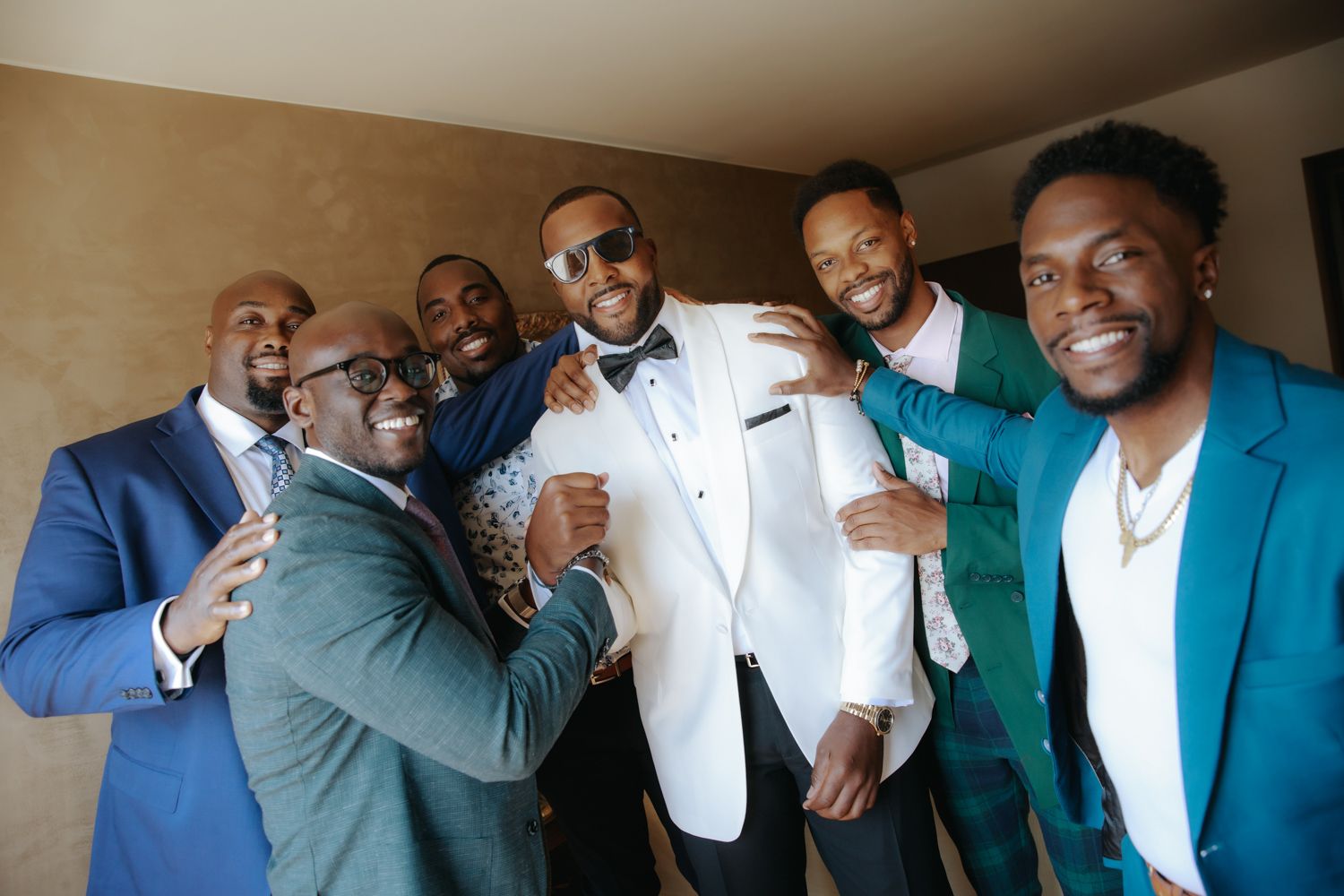 Groom in white tuxedo celebrating with groomsmen in blue and green suits at a wedding.