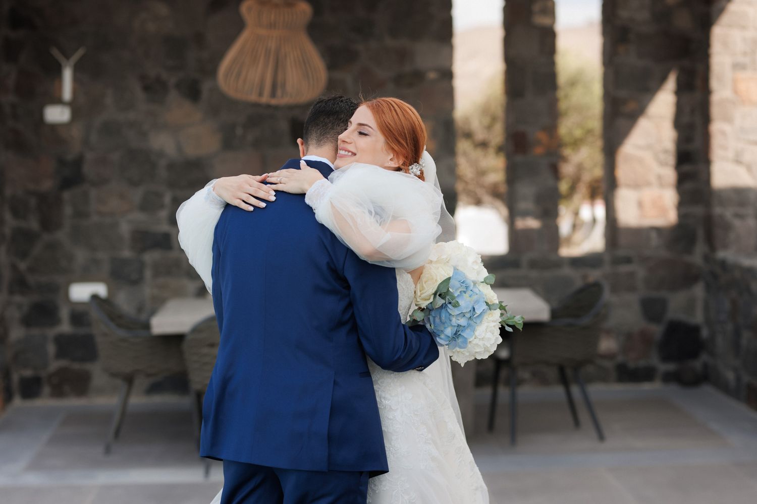 Bride with red hair in white gown embracing groom in blue suit holding blue hydrangea bouquet.