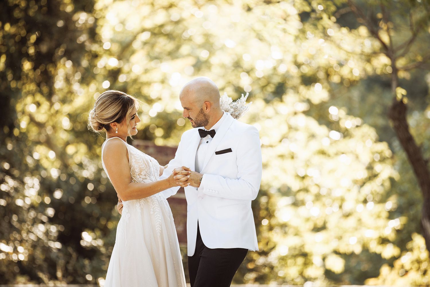 Wedding couple shares a tender moment in a sun-dappled forest setting wearing white formal attire.