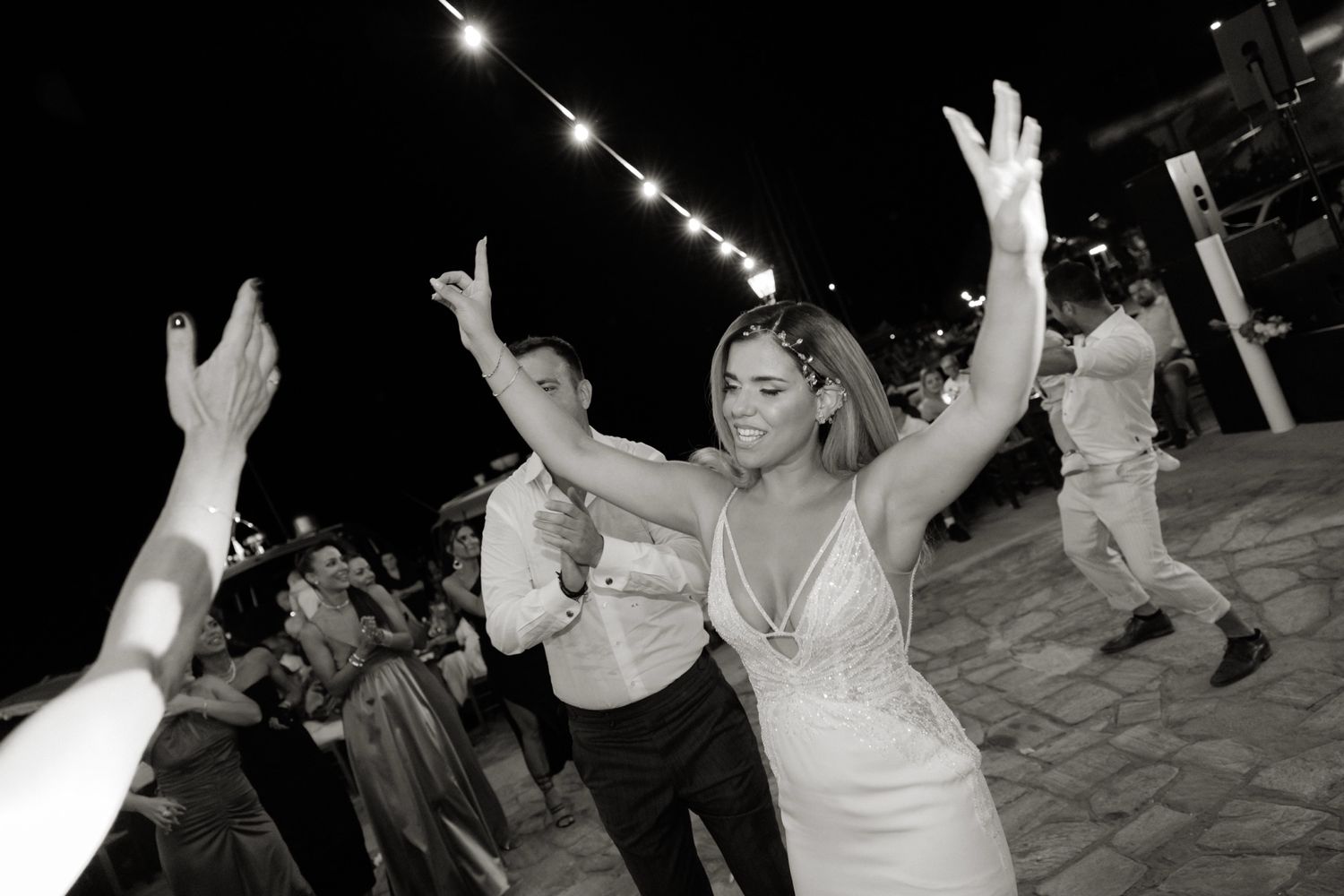 Bride dancing joyfully with raised arms at outdoor wedding reception in black and white photo.