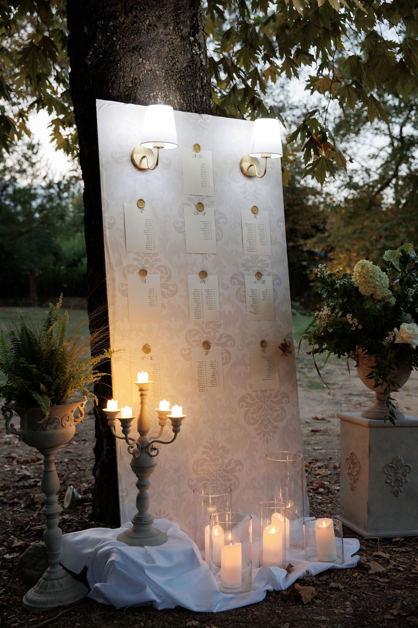Romantic outdoor wedding table plan board illuminated by candlelight with white floral decor and candelabra at dusk.