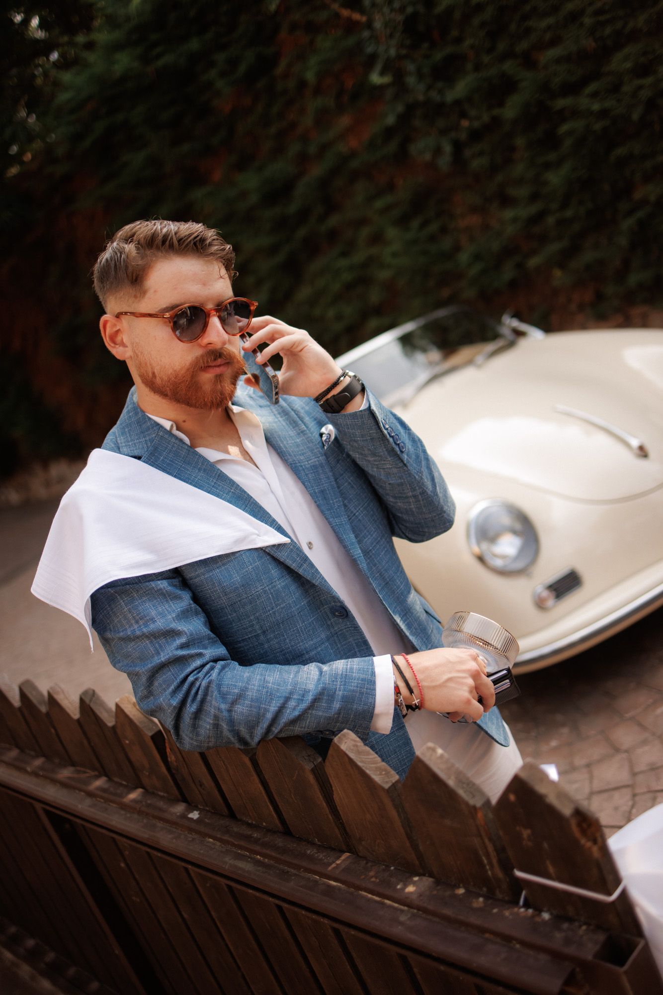 Stylish man in blue blazer and sunglasses talking on phone beside a vintage white Porsche on a sunny day.
