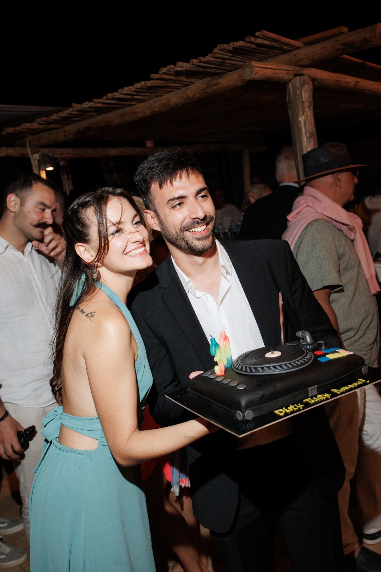 Smiling couple posing together holding a DJ turntable themed birthday cake at night.