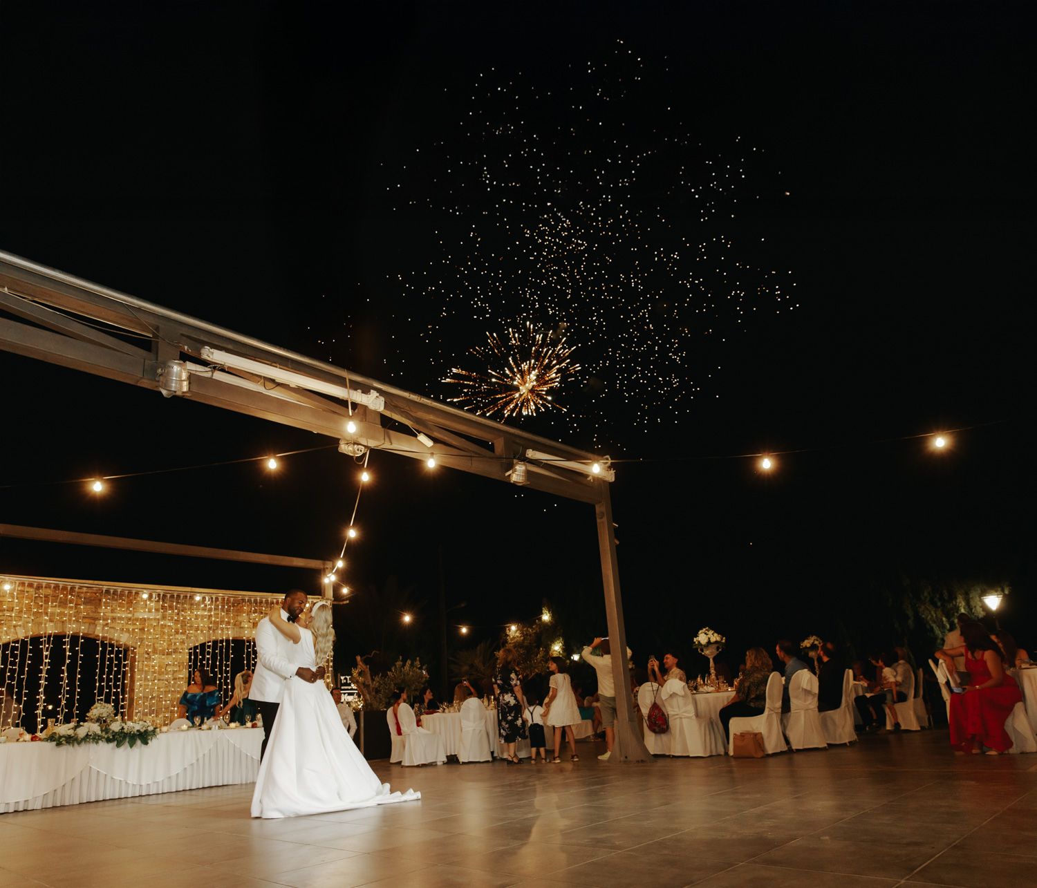 Bride and groom share first dance under string lights with sparkling fireworks at an outdoor wedding reception.