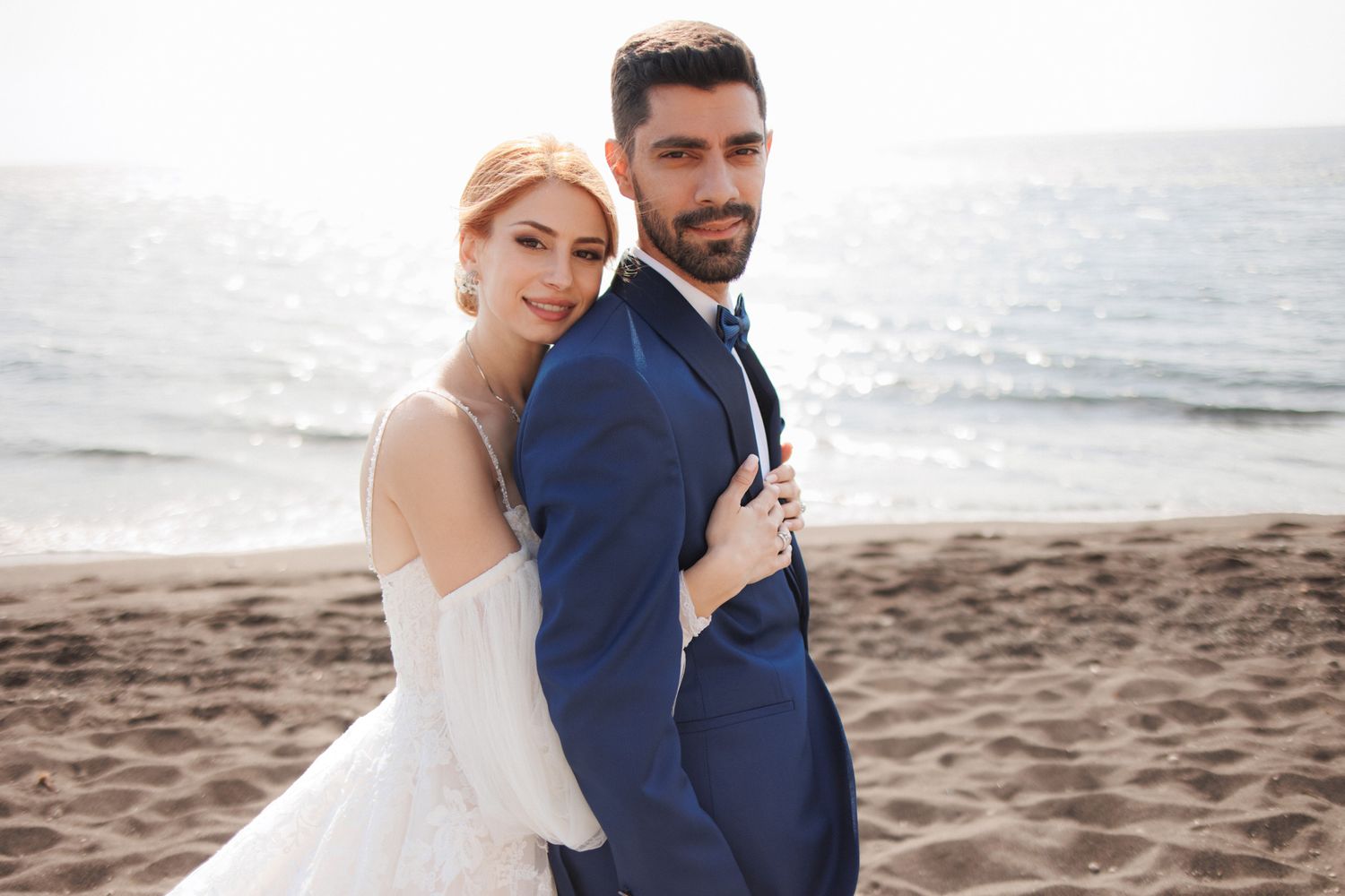 Bride in off-shoulder gown embracing groom in navy suit on a sandy beach at golden hour.