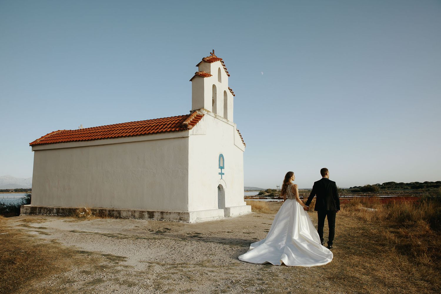 A couple walks away from a small white chapel with red roof tiles on a sunny day in a rural landscape.