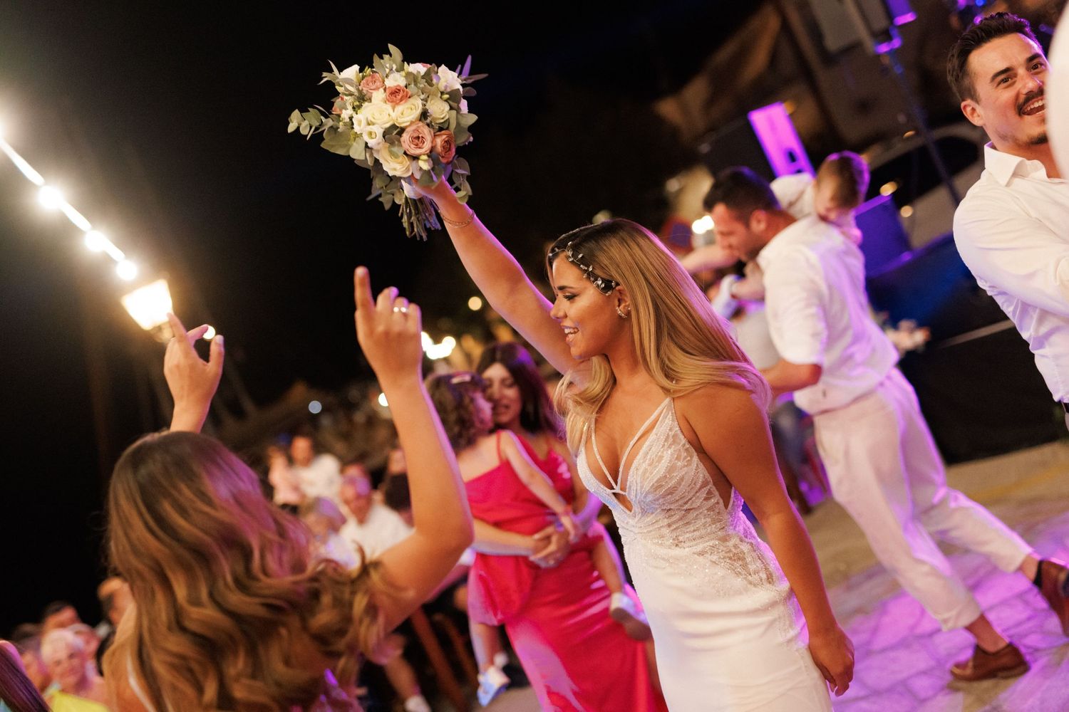 Bride in white gown tossing bouquet to guests during lively outdoor wedding reception at night.