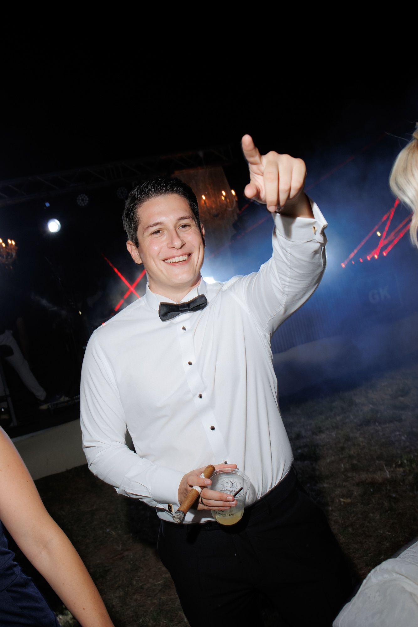 Smiling groom in white shirt and bow tie pointing while holding drink at wedding.