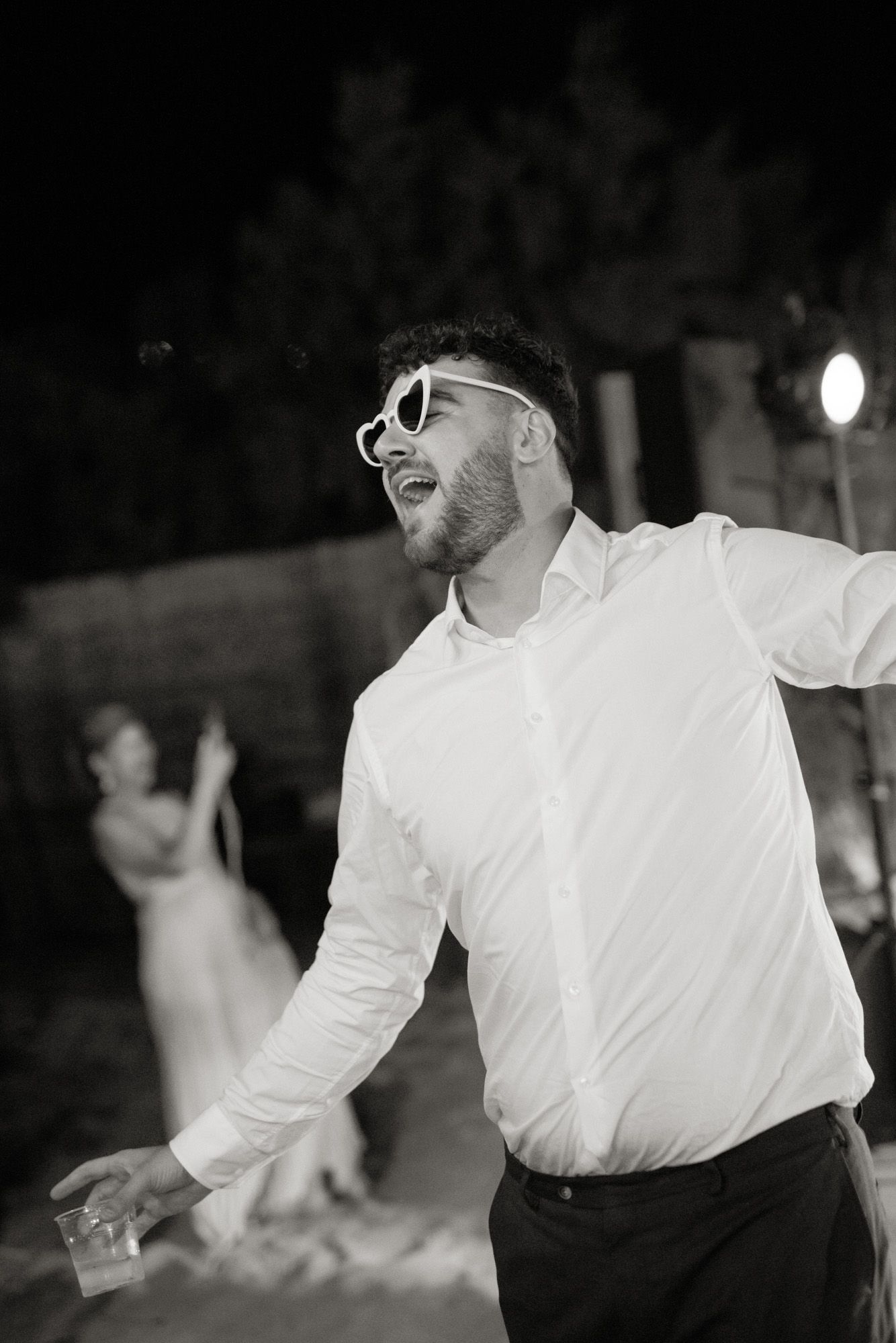 Man in white shirt and sunglasses dancing joyfully at an outdoor nighttime wedding reception.