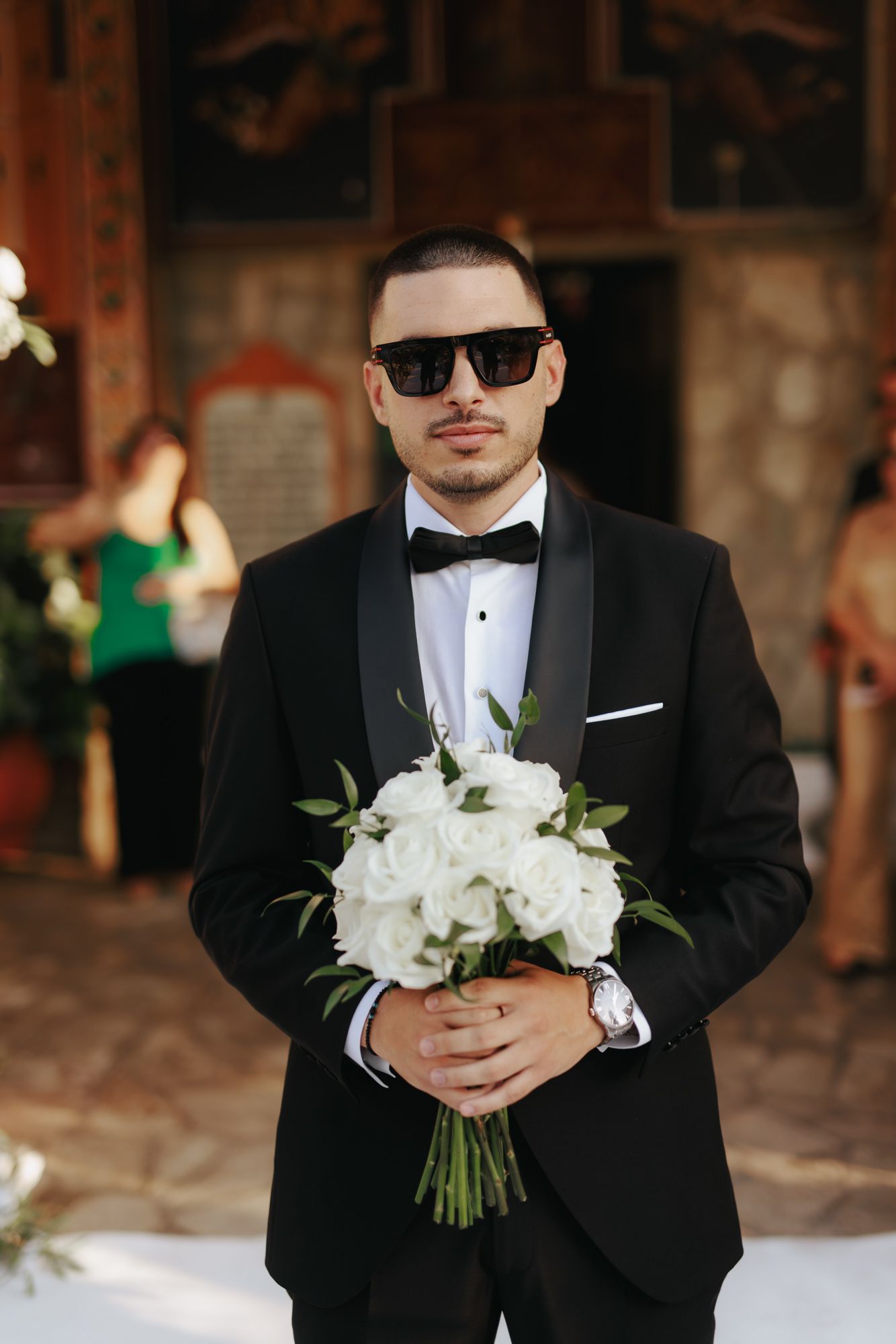 Elegant groom in black tuxedo and sunglasses holding white rose bridal bouquet at outdoor wedding ceremony.