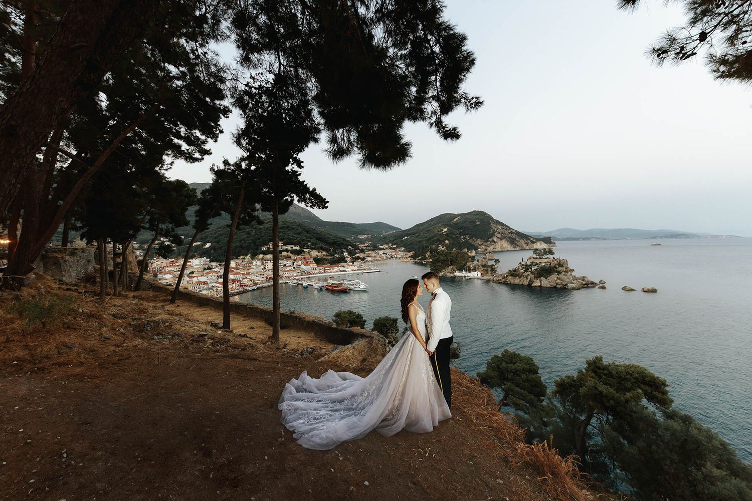 A couple stands on a cliff overlooking a Mediterranean coastal bay with pine trees framing the scenic view.