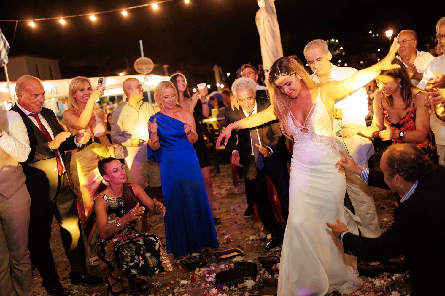 Bride in white gown dancing with guests at an outdoor wedding reception under warm lights.