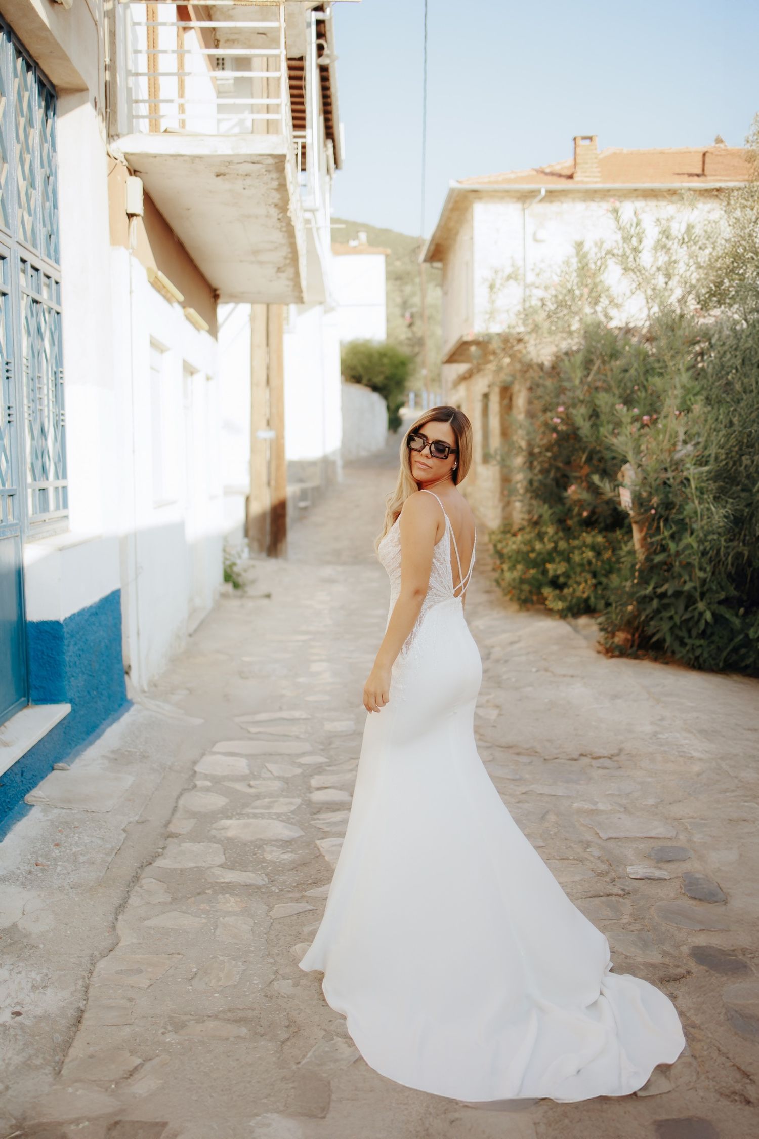 Bride in elegant white mermaid wedding dress with train walking along a sunny Mediterranean alley.
