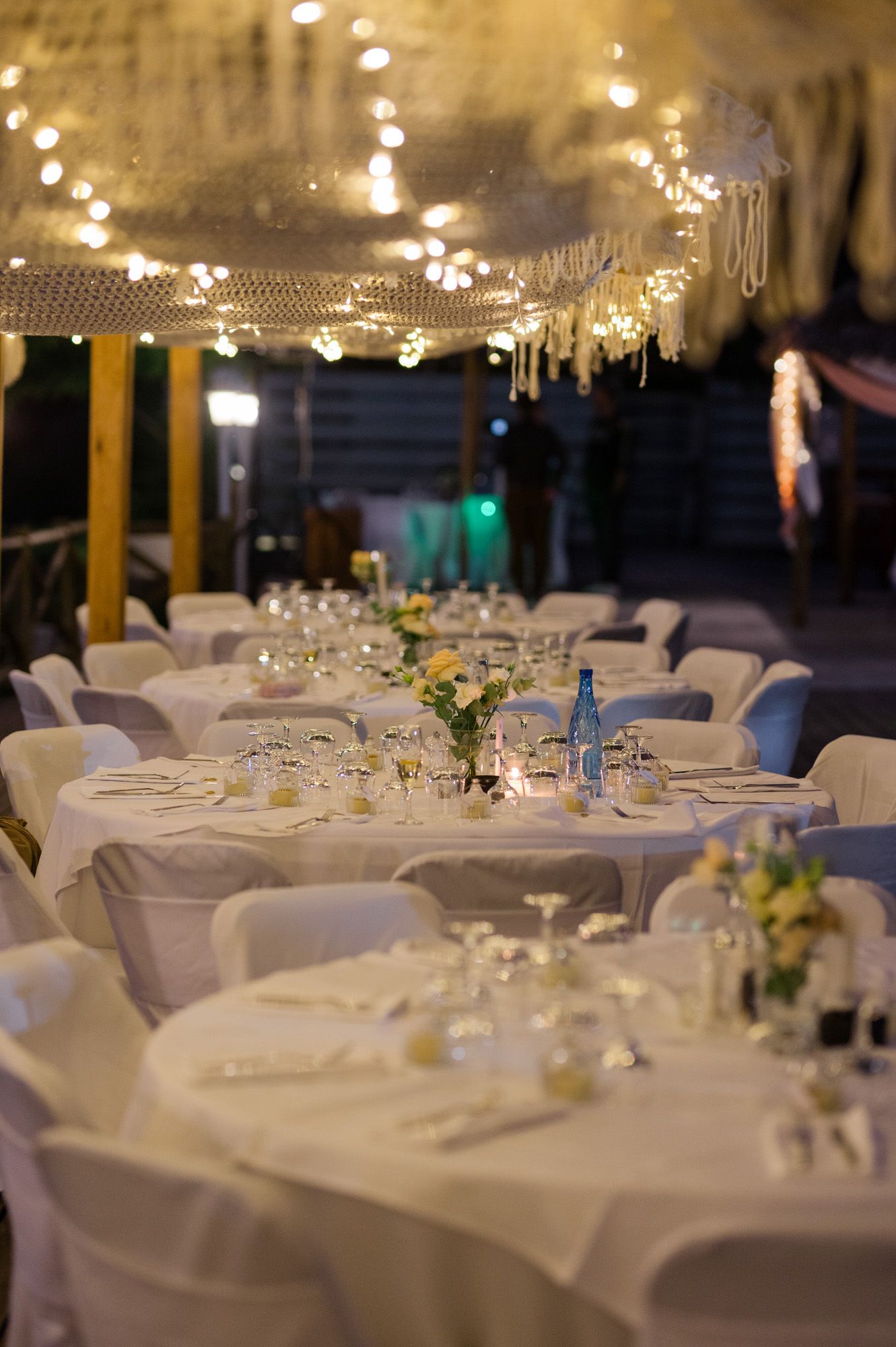 Elegant wedding reception tables with white linens, floral centerpieces, and string lights overhead.