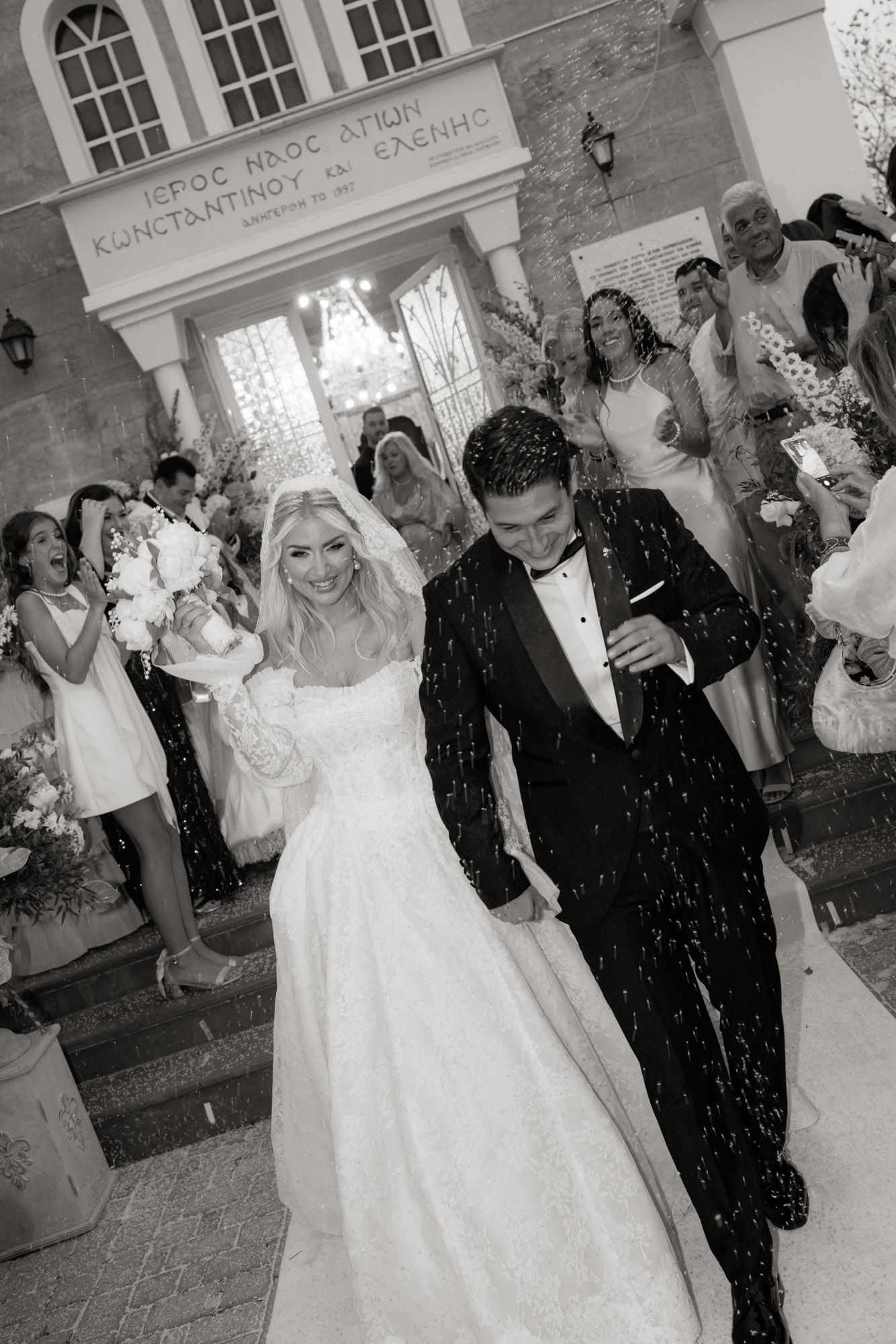 Newlyweds walk through rice shower outside Greek Orthodox church of Saints Constantine and Helen in black and white.