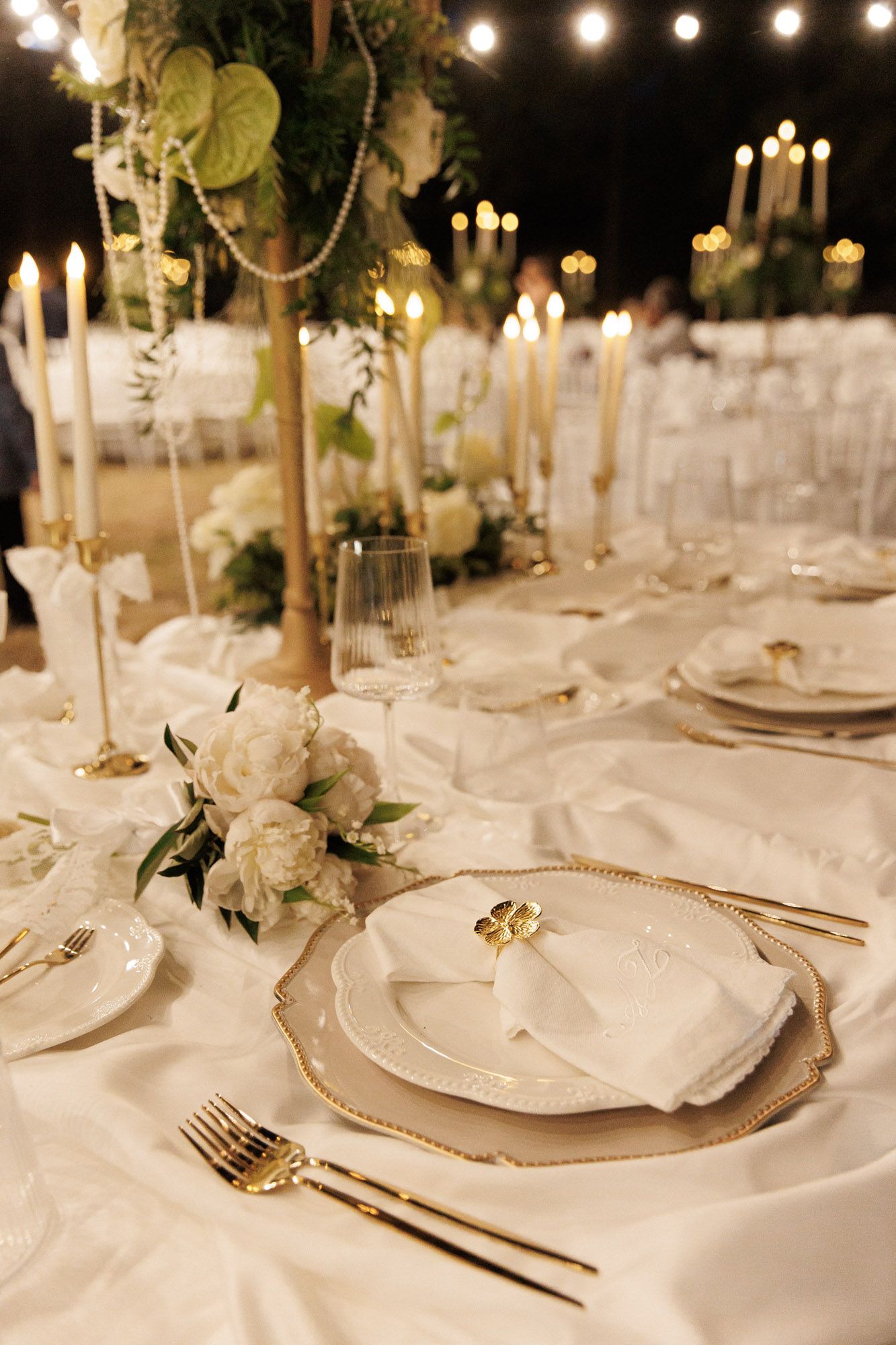 Elegant outdoor wedding reception tables set with white linens, gold candelabras, and lush floral centerpieces at night.