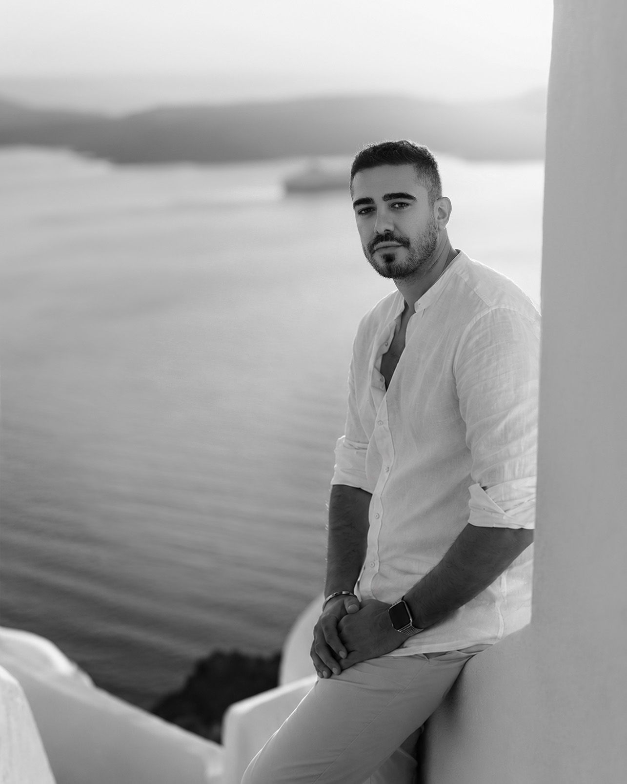 Man in white linen shirt posing against Santorini sea view in black and white portrait photo.