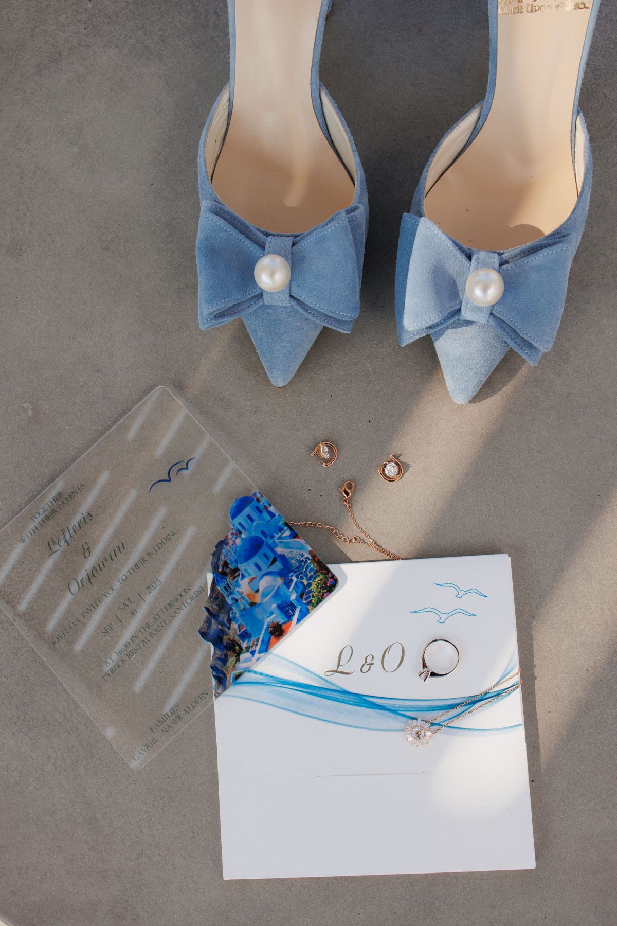Blue suede bow heels with pearl detail alongside gold jewelry and wedding invitation suite on gray surface.