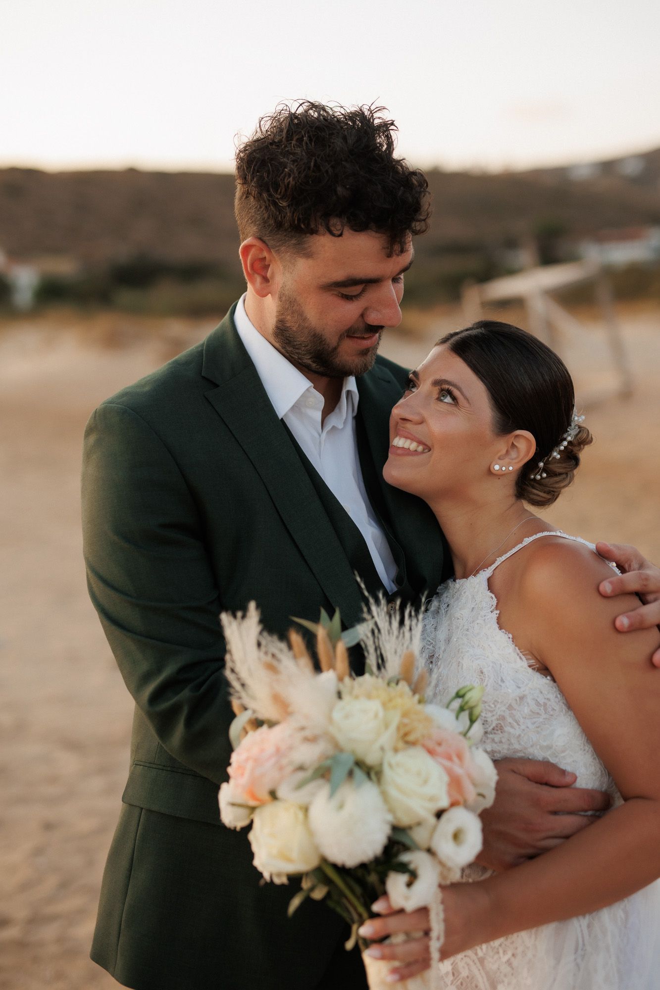 Bride smiles up at groom as they embrace on sandy beach at golden sunset.