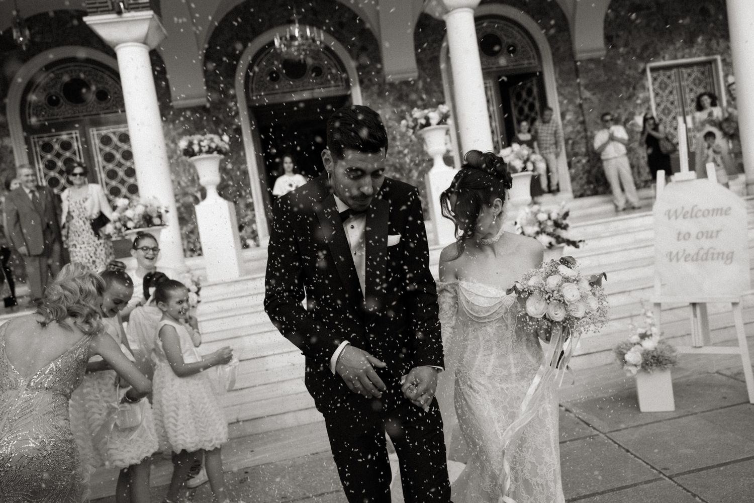 Black and white photo of newlyweds exiting church in confetti shower, bride holding bouquet, Welcome to our Wedding sign.