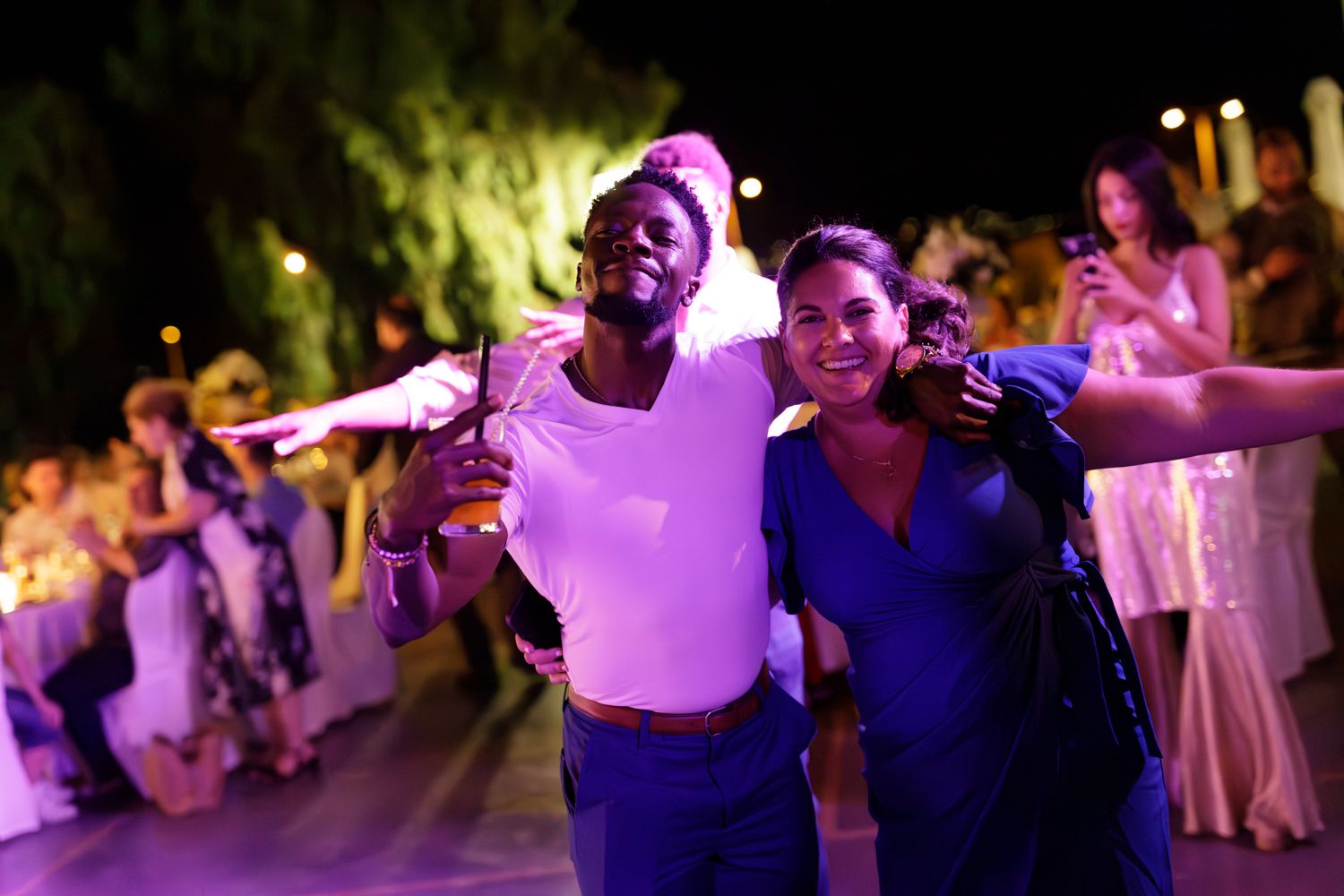 Two joyful friends pose together at an outdoor evening party, with purple lights and guests dancing in the background.