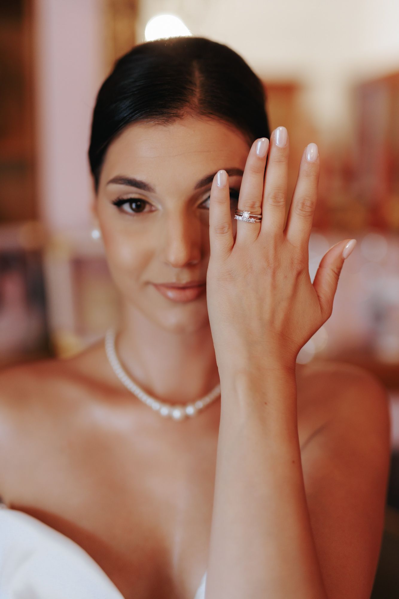 Bride in white dress and pearl necklace showing diamond engagement ring on her finger while posing elegantly.