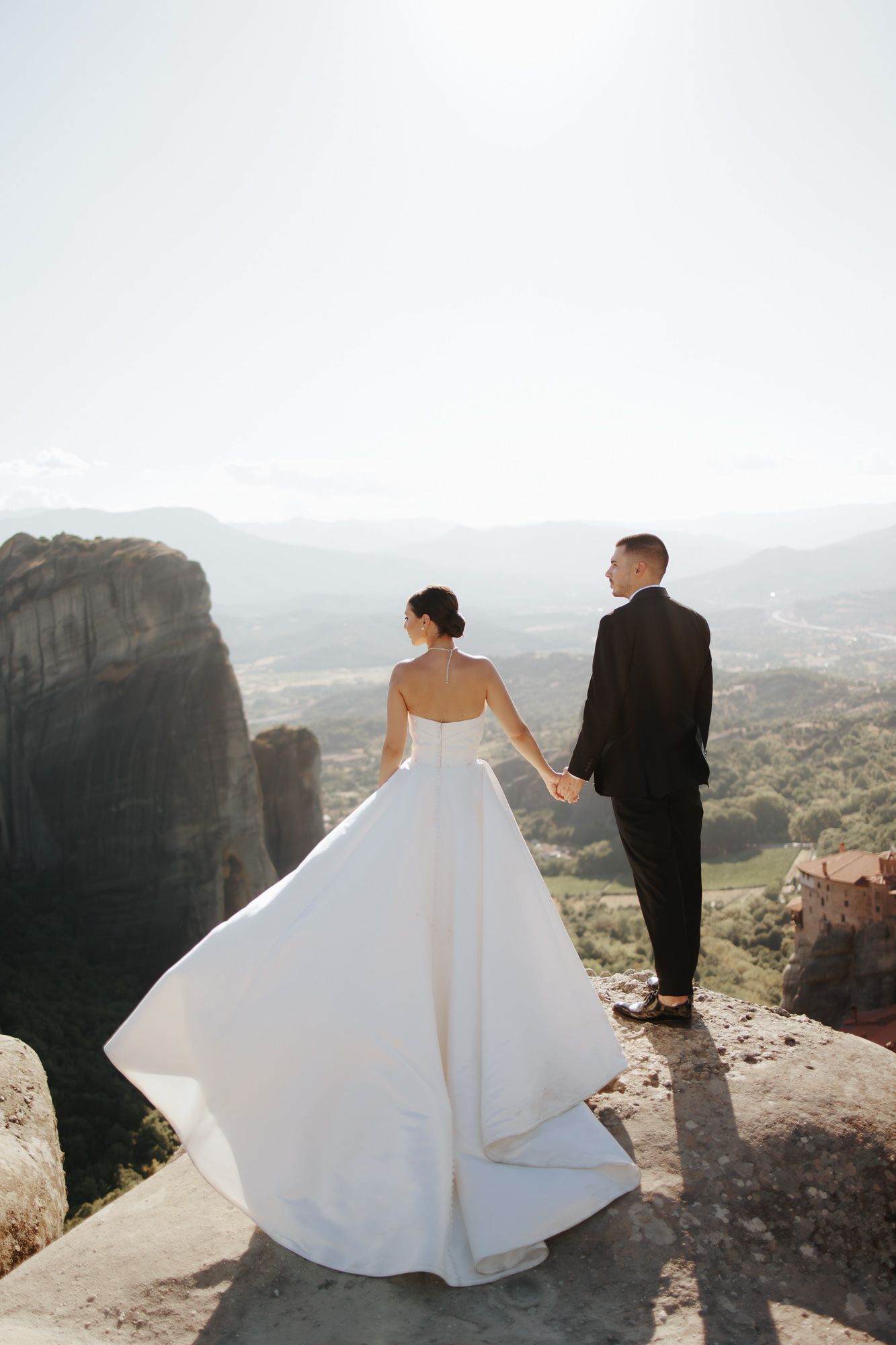 Bride and groom holding hands on a cliff edge at Meteora, Greece, with stunning mountain valley views.