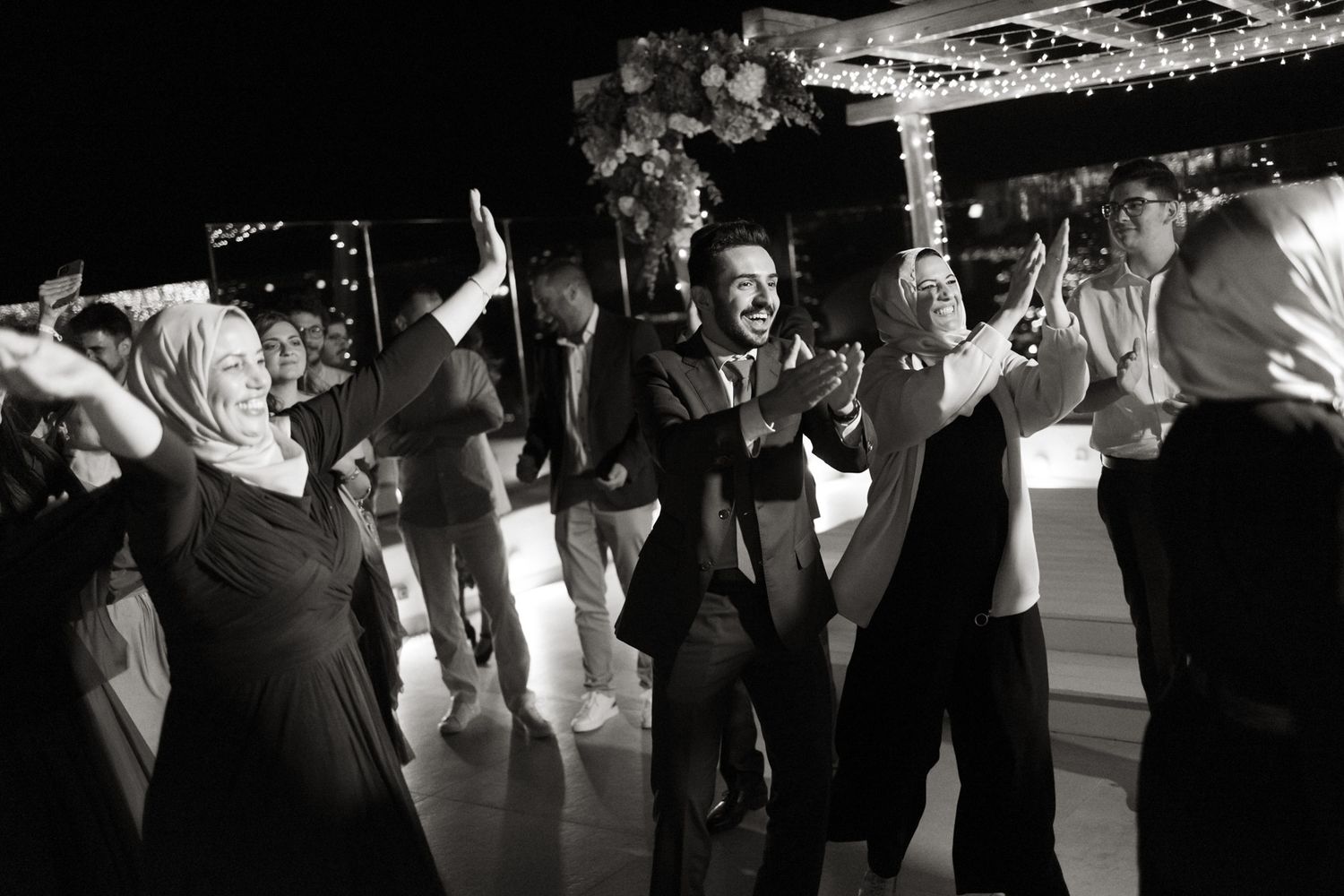 Wedding guests dance joyfully with hands raised at lively black and white evening reception.