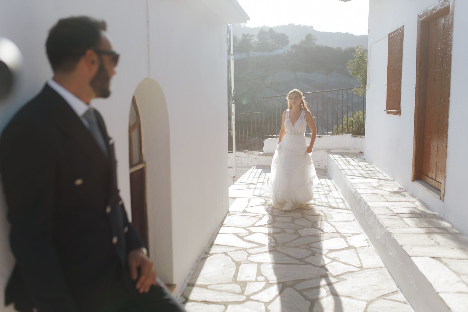 Groom in dark suit watches bride in white wedding gown approach along a sunlit Greek pathway.