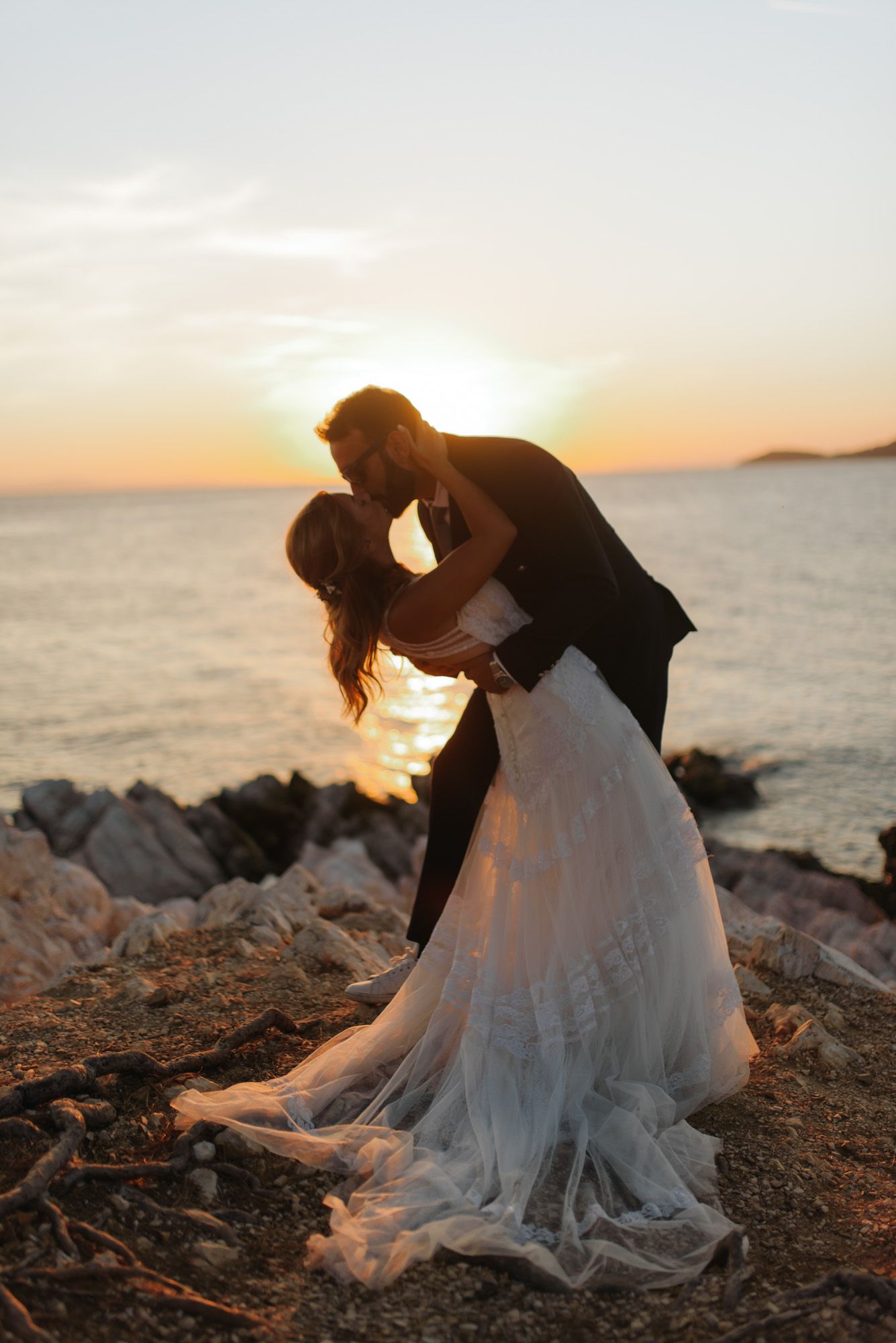 Bride and groom sharing a romantic dip kiss on rocky beach during breathtaking golden sunset.