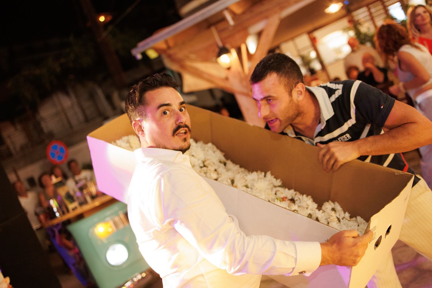 Two men playfully pose with large cardboard box filled with white packing peanuts at wedding.