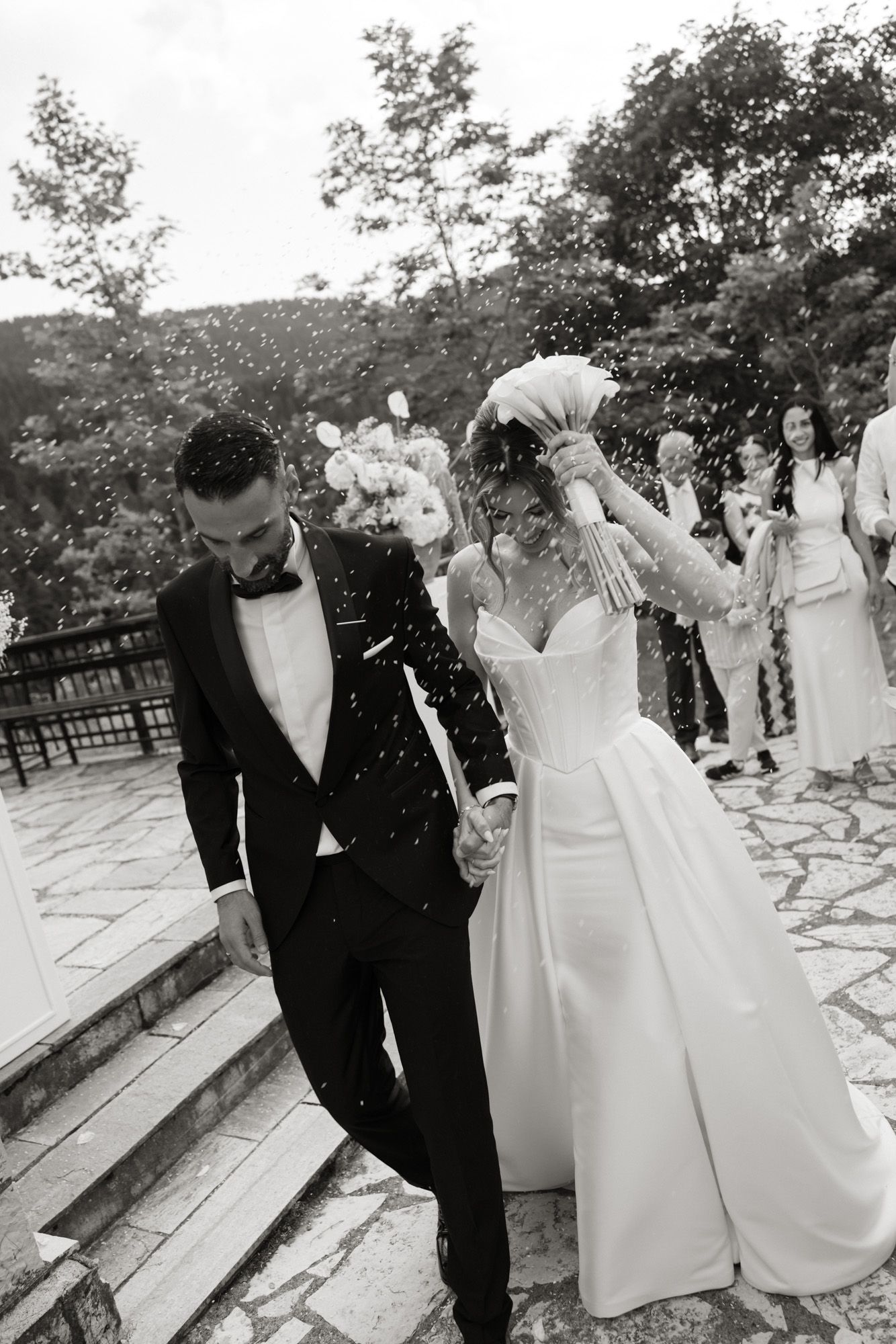 Newlyweds walk hand-in-hand through rice shower after outdoor wedding ceremony in black and white photo.