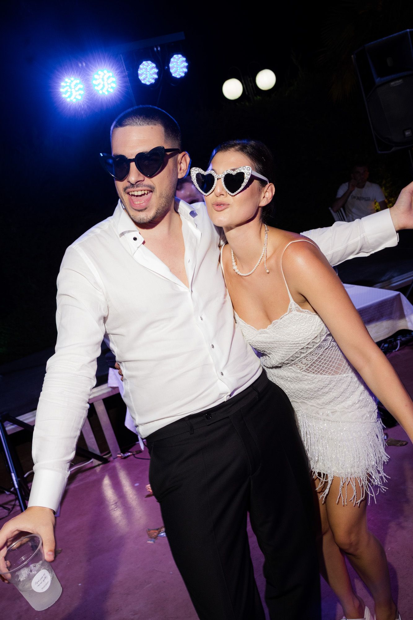 Couple in matching white outfits and heart-shaped sunglasses dancing at a nightclub event.