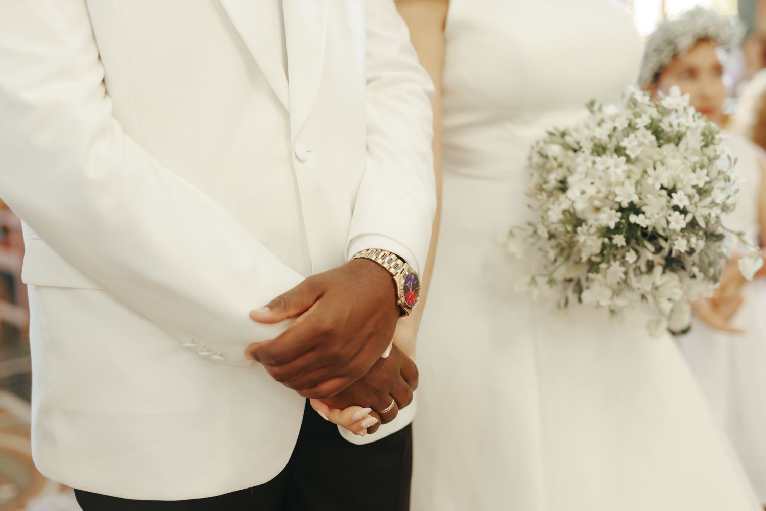 Bride and groom holding hands at wedding ceremony, groom in white suit, bride holding white baby's breath bouquet.