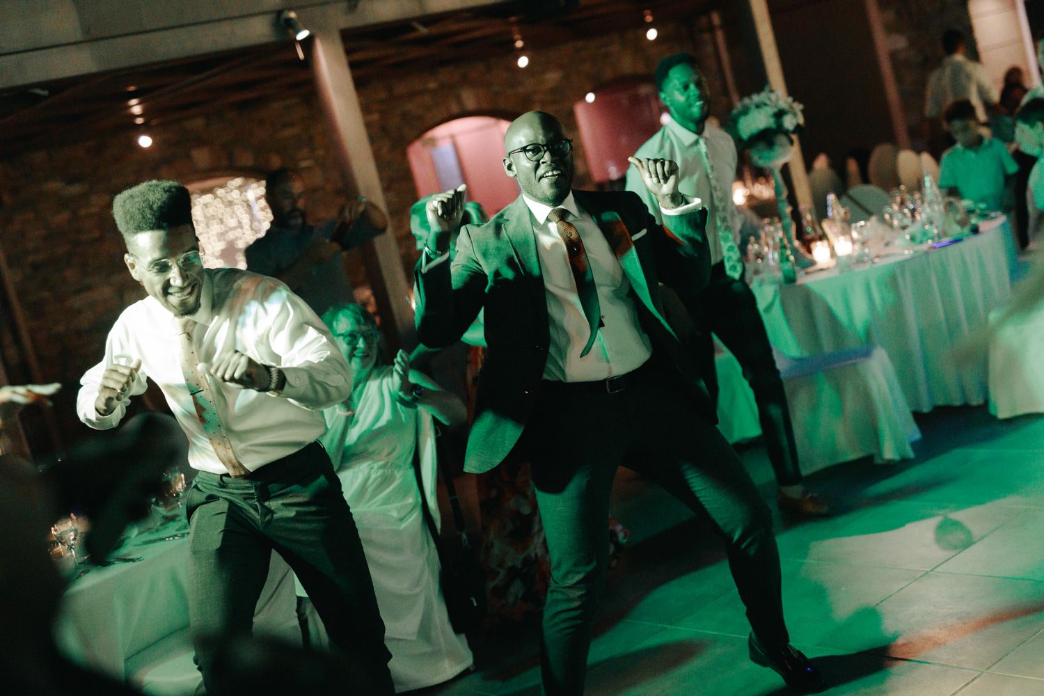 Men in formal attire dancing energetically at an elegant wedding reception with green lighting.