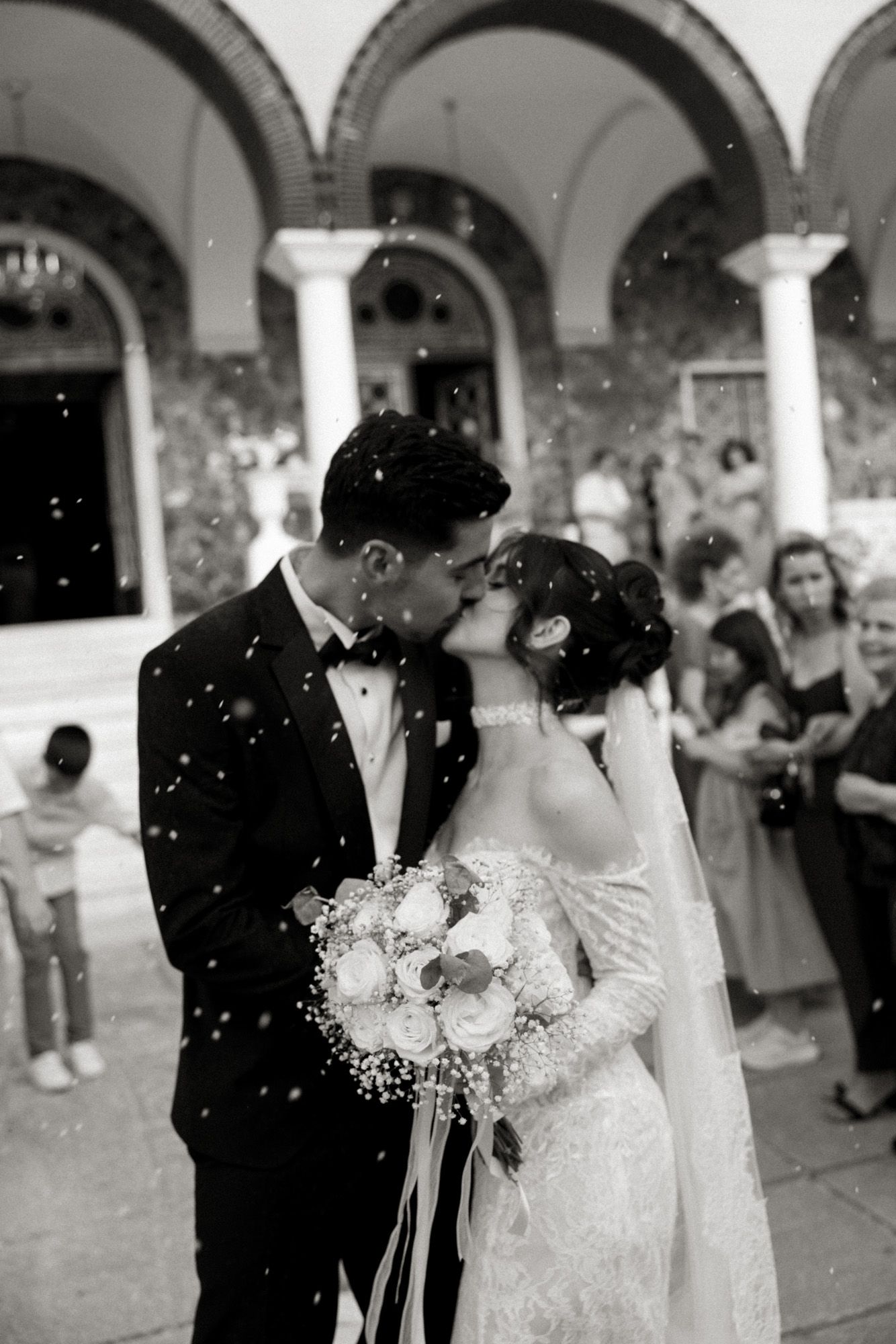 Black and white photo of a bride and groom sharing a kiss outside a church while confetti falls around them.