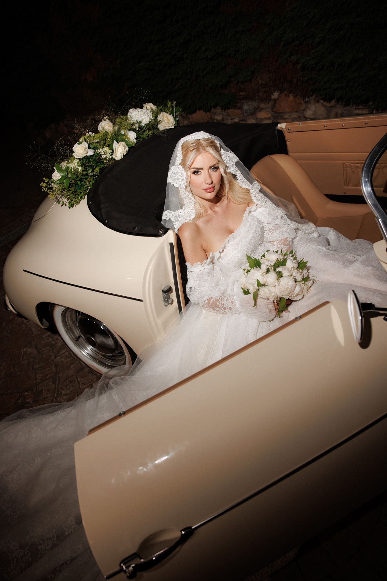 Elegant bride in lace wedding gown and veil seated in vintage cream convertible car at night holding white bouquet.