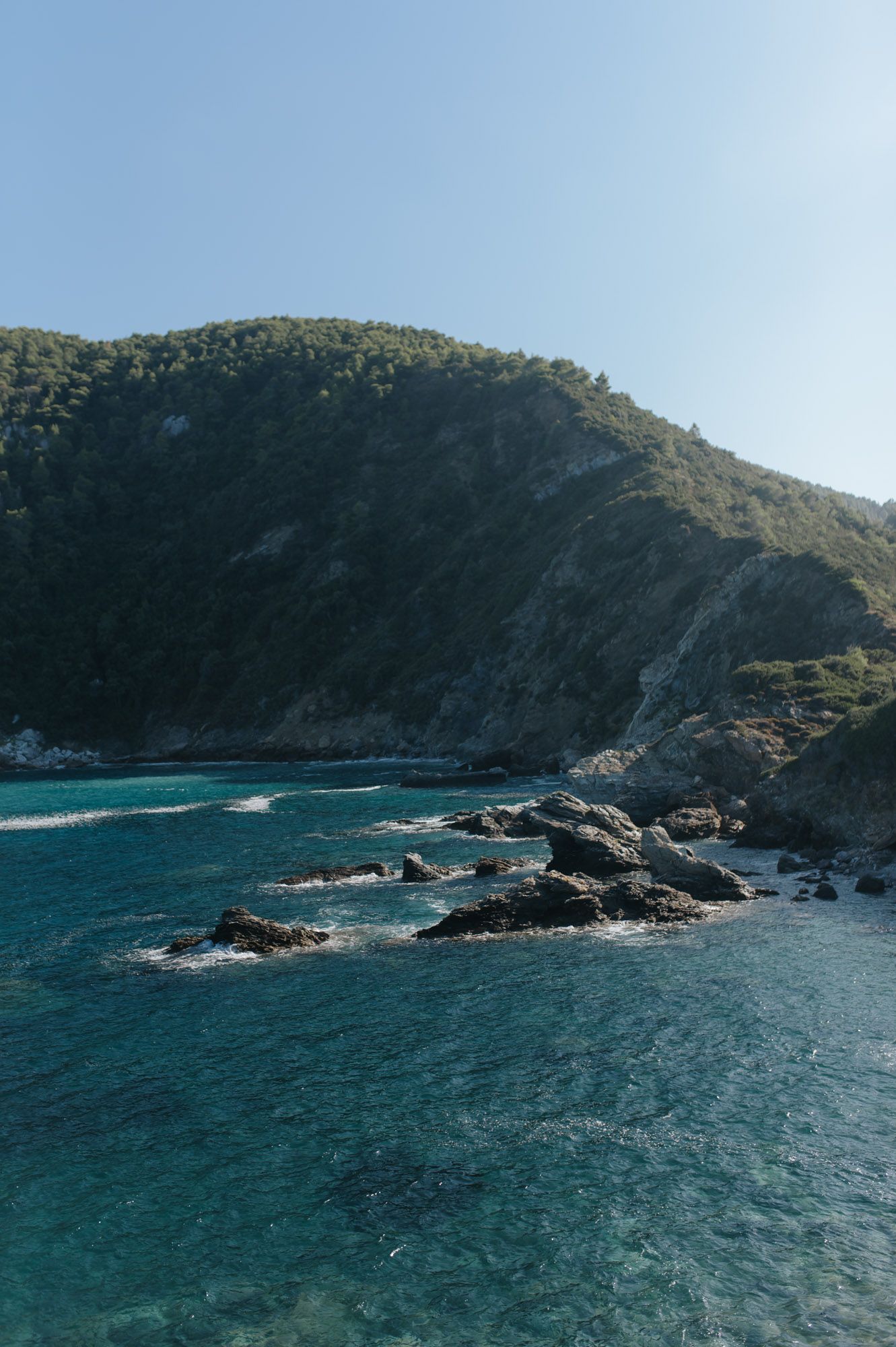 Stunning turquoise Mediterranean sea with rocky coastline and lush green hillside under a clear blue sky.
