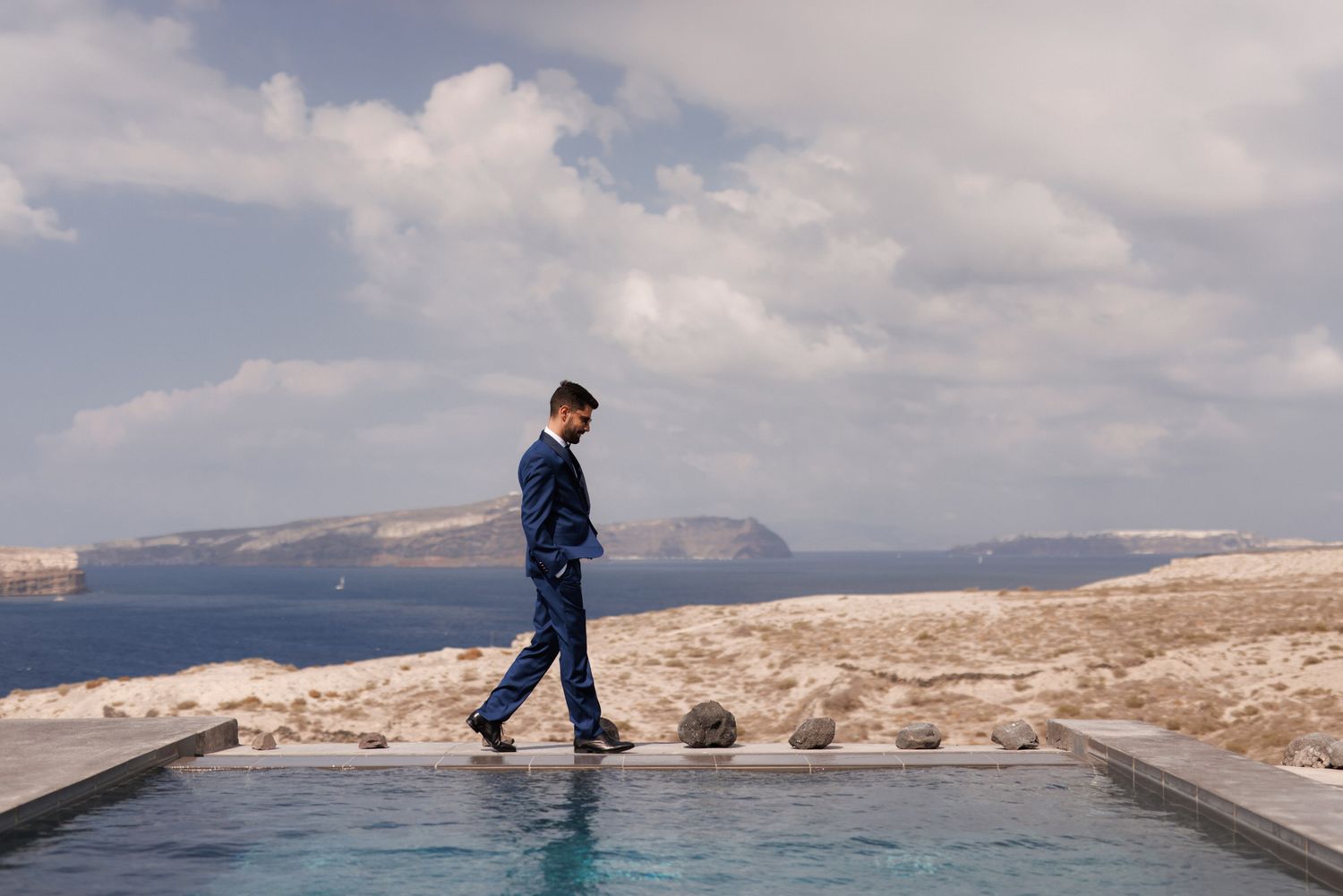Groom in navy suit walking along infinity pool edge with scenic Santorini landscape behind.