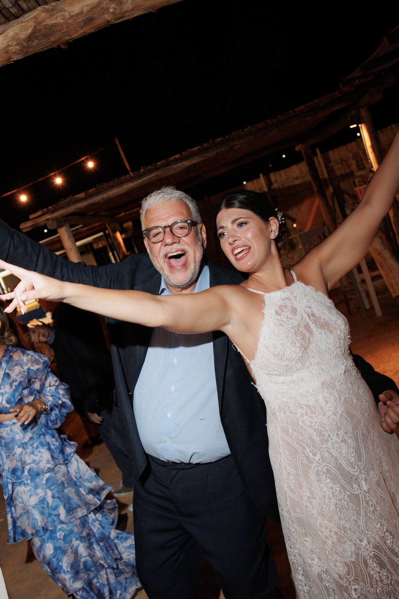 Bride in halter lace gown dances joyfully with older man in glasses at wedding reception.