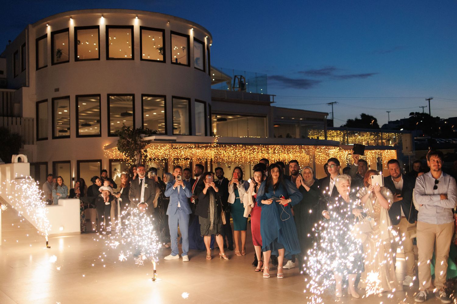 Wedding guests celebrate outdoors at night with sparklers in front of beautifully lit venue.