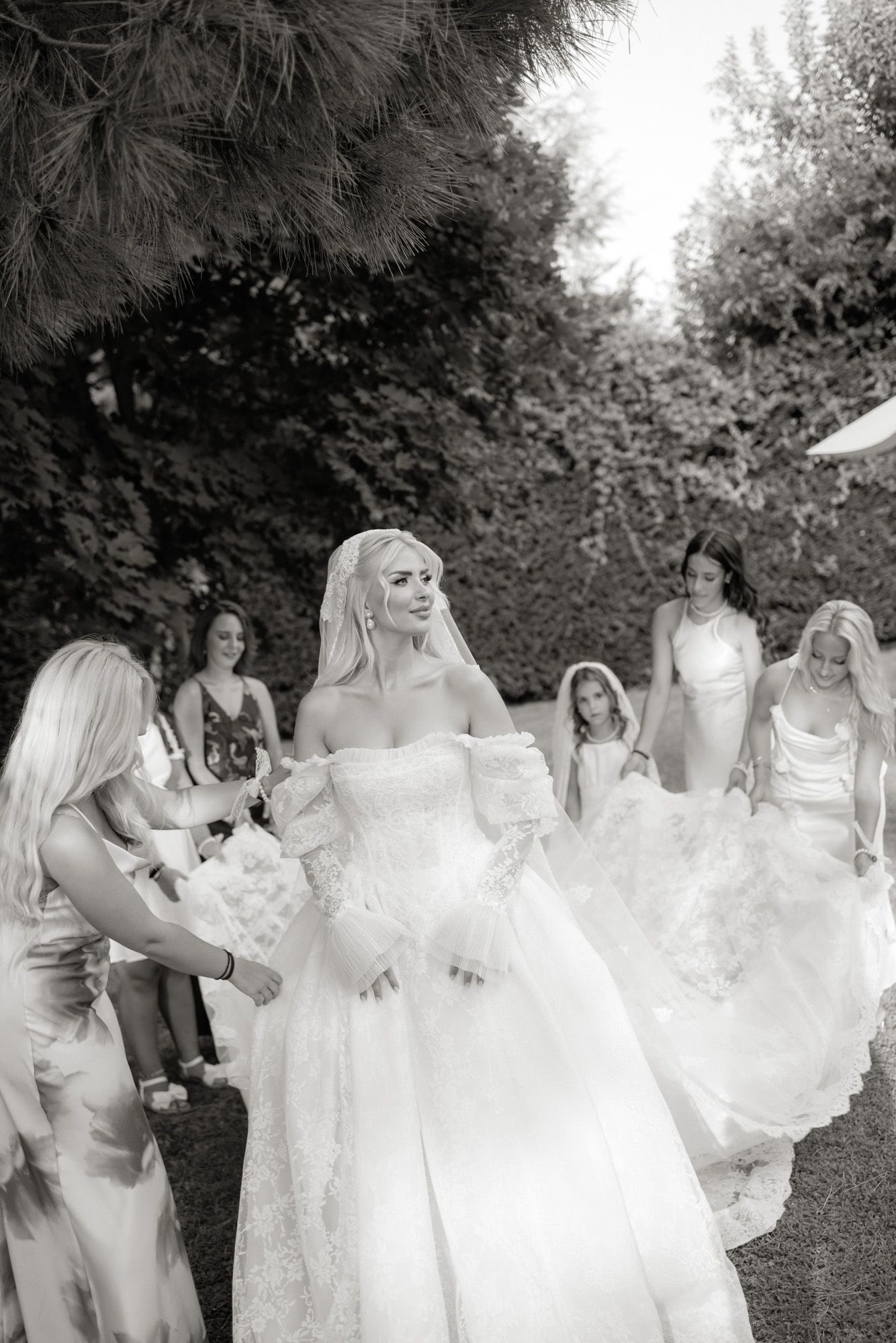 Bride in off-shoulder lace gown walks with bridesmaids and flower girl in black and white garden wedding photo.