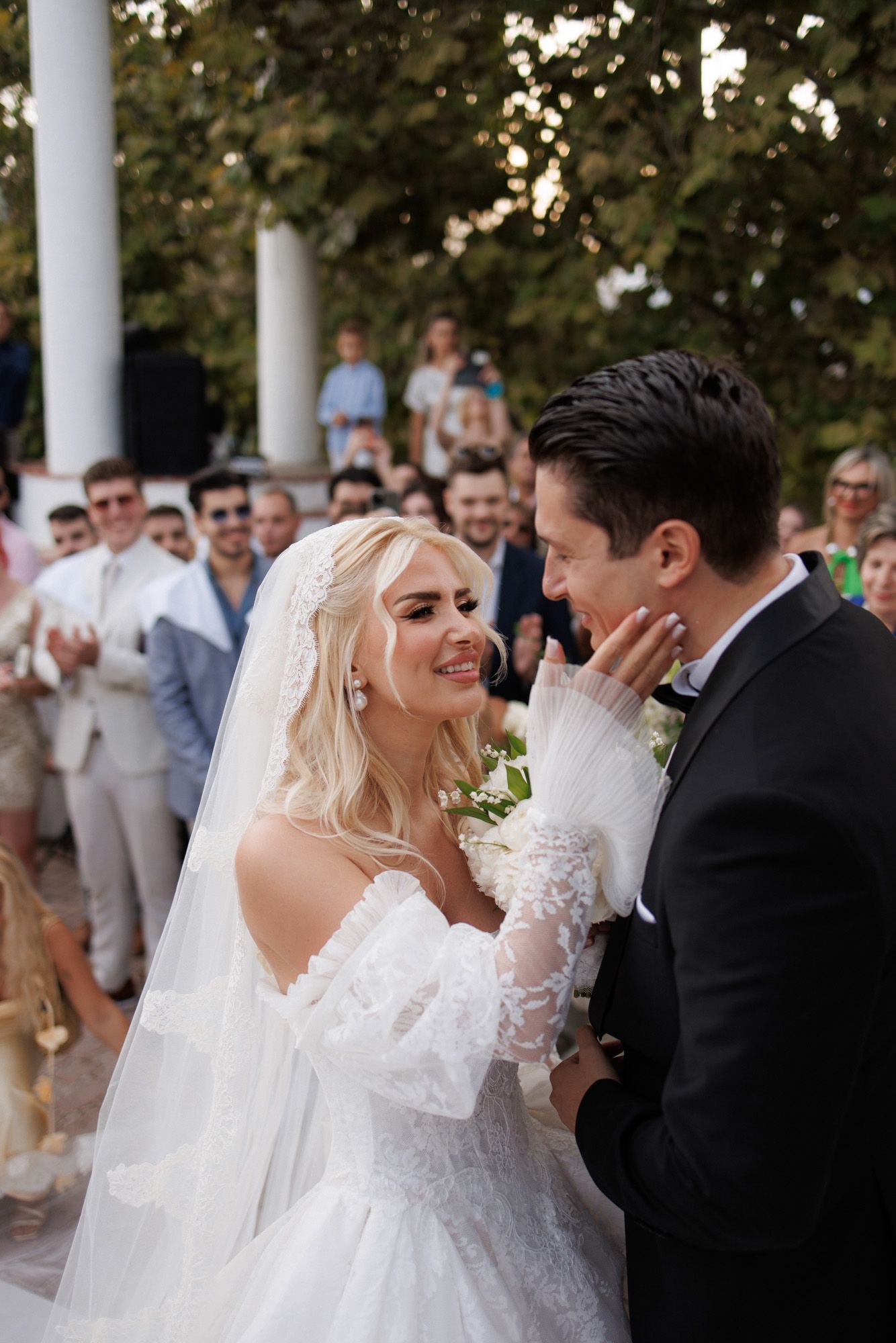 Emotional bride and groom share an intimate moment surrounded by cheering wedding guests outdoors.