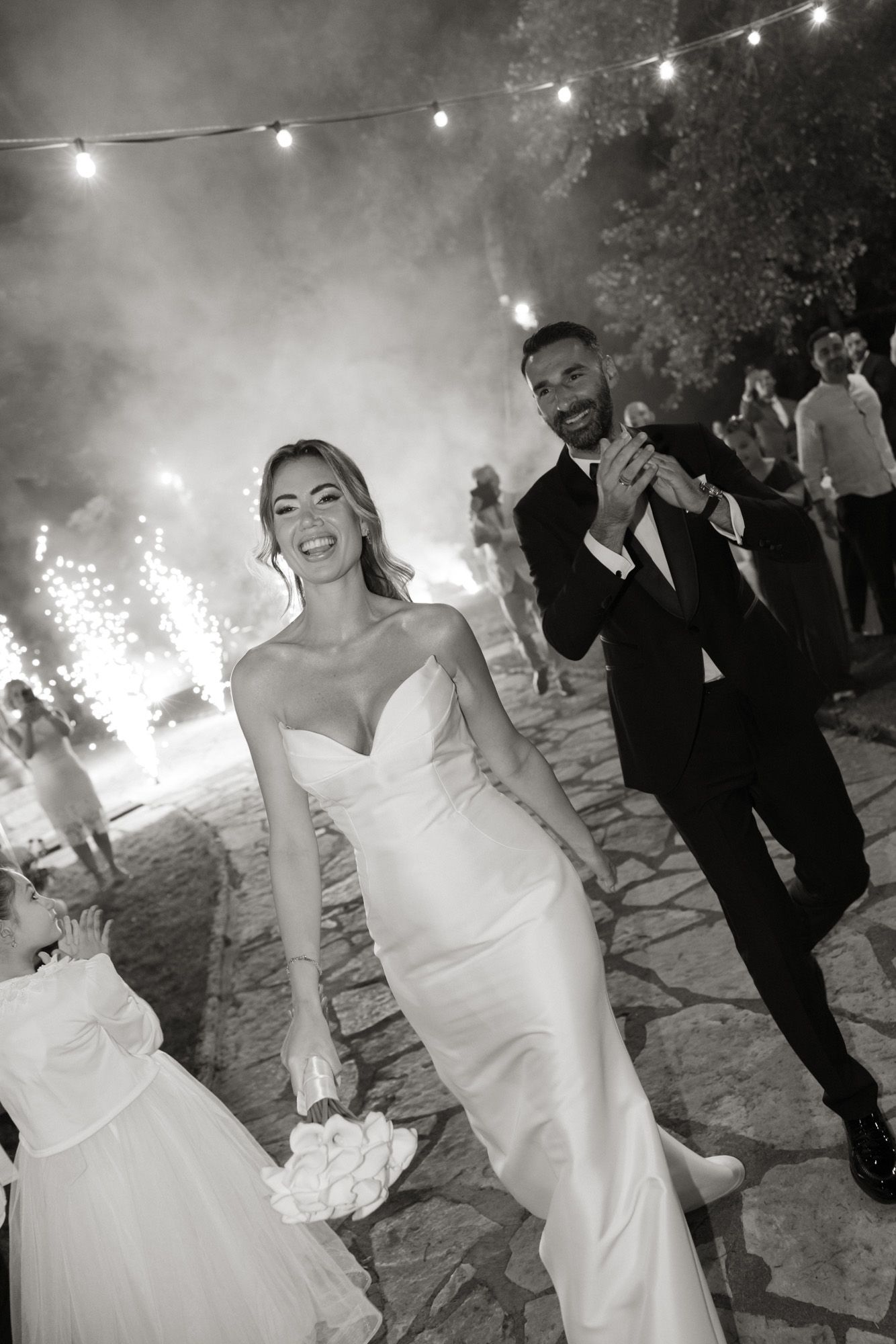 Smiling bride and groom walk through sparklers and smoke at nighttime wedding celebration in black and white.