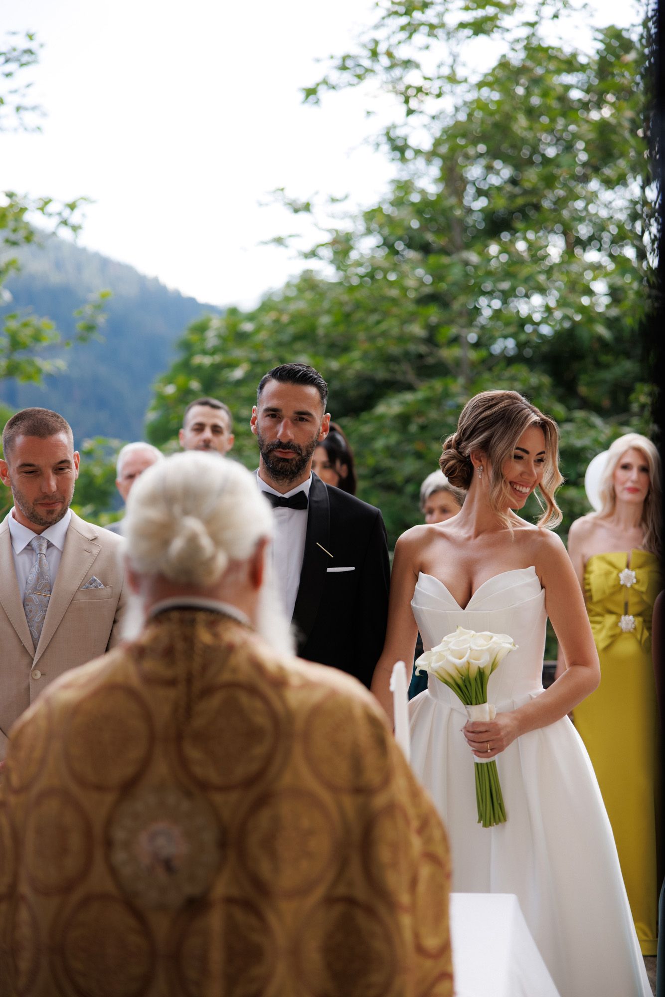 Bride holding white calla lily bouquet stands before officiant during outdoor wedding ceremony in mountain setting.