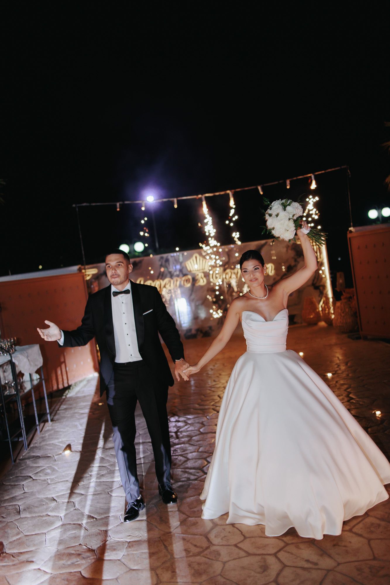 A joyful bride in a white ballgown and groom in a tuxedo celebrate their wedding reception outdoors at night with sparkling lights.