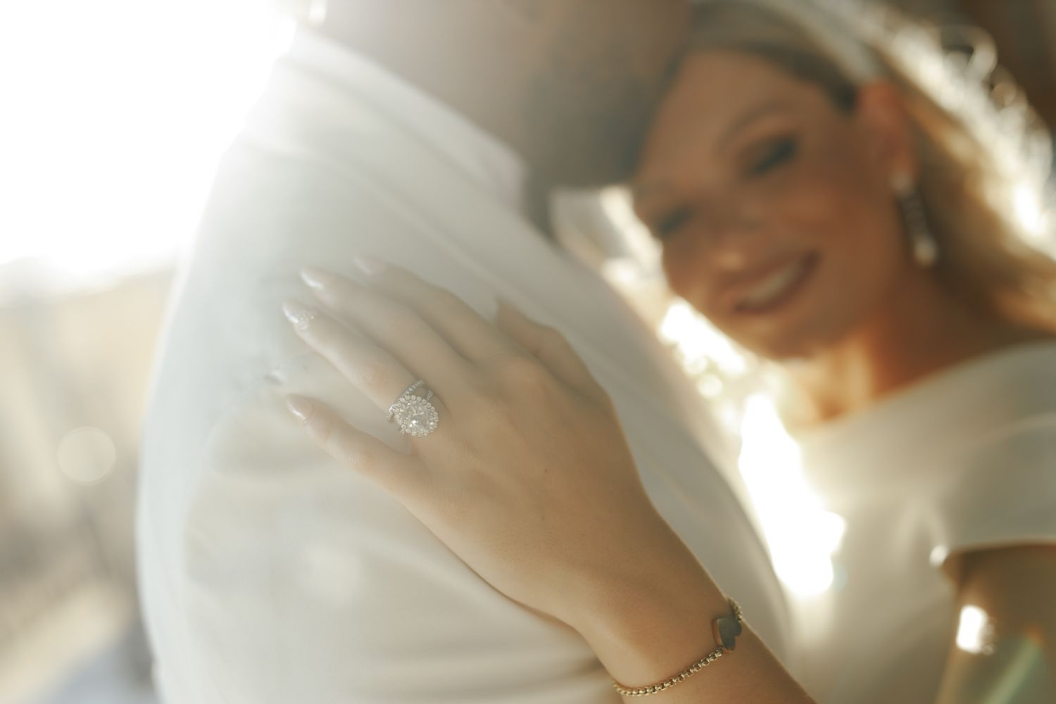 Smiling bride showing diamond engagement ring while embracing groom in soft golden sunlight.