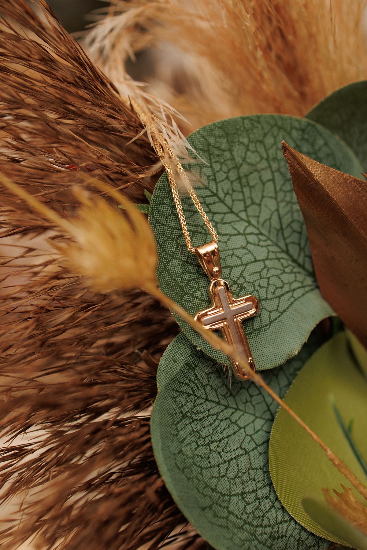 Gold cross pendant necklace displayed on green leaves with dried pampas grass in warm earthy tones.